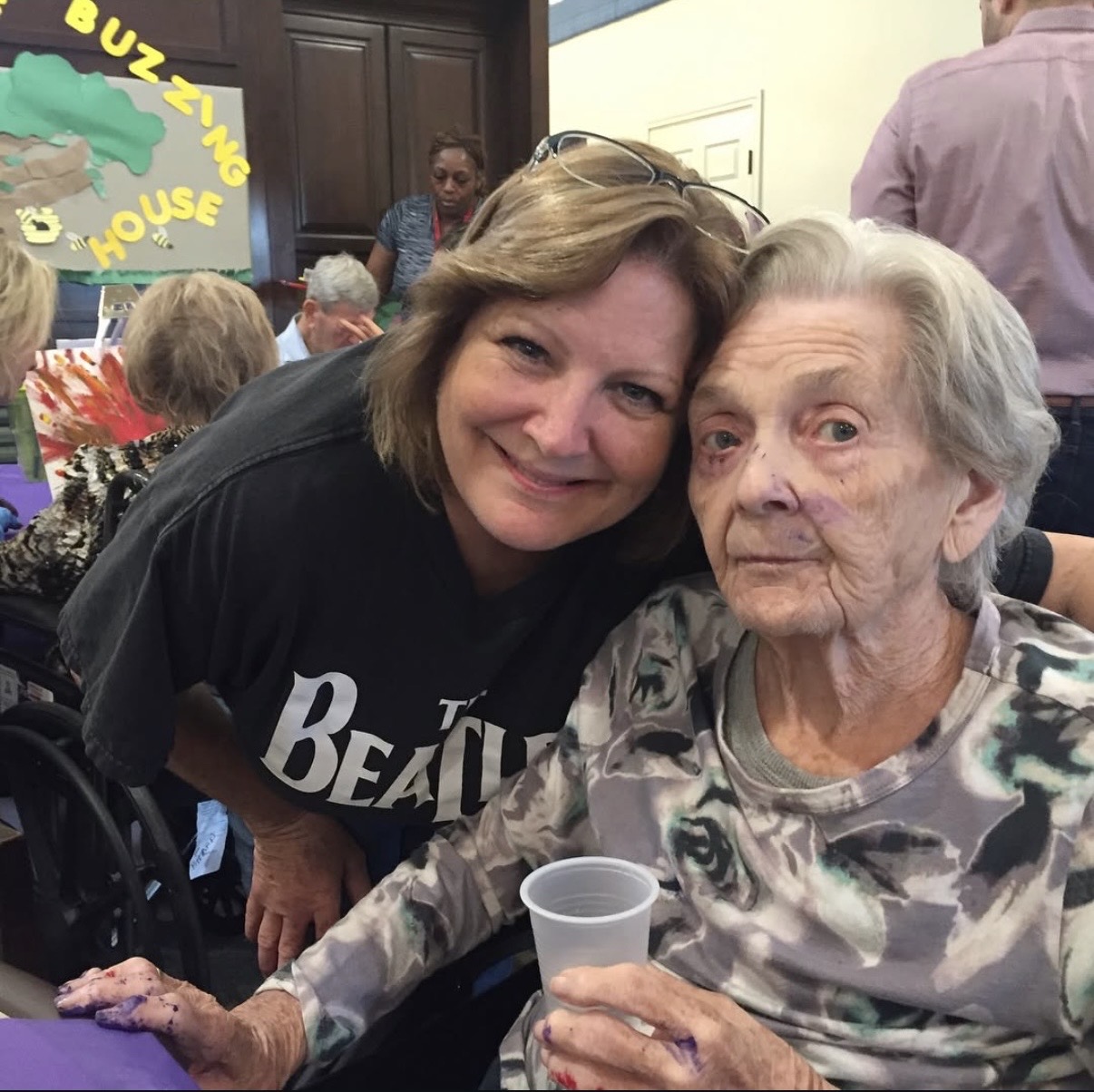 Two women, one elderly and one middle-aged, smiling and sitting together at an event, with a crowd and a sign in the background.
