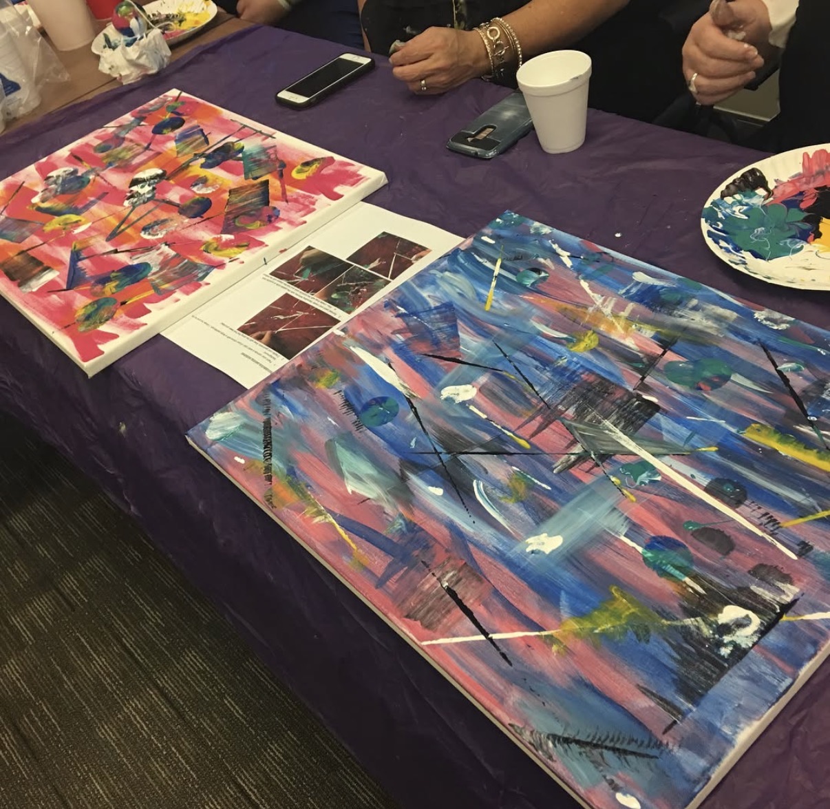 Two colorful abstract paintings on a table with people, phones, and cups nearby.