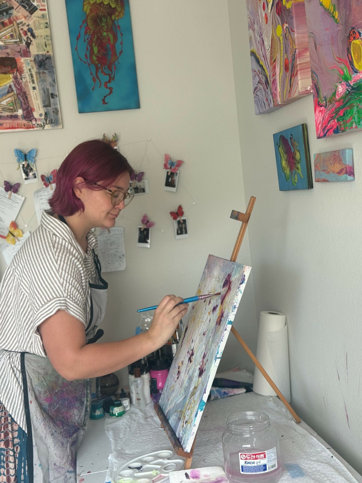 Woman with glasses and short red hair painting on canvas in art studio, surrounded by colorful artwork on walls.