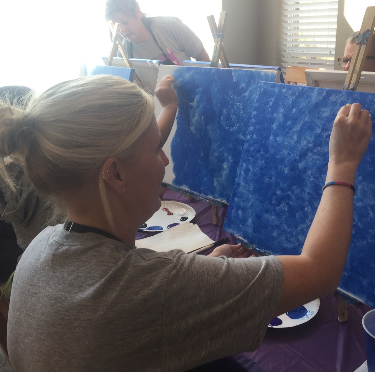 Person painting a blue canvas with a brush, seated at a table with painting supplies.
