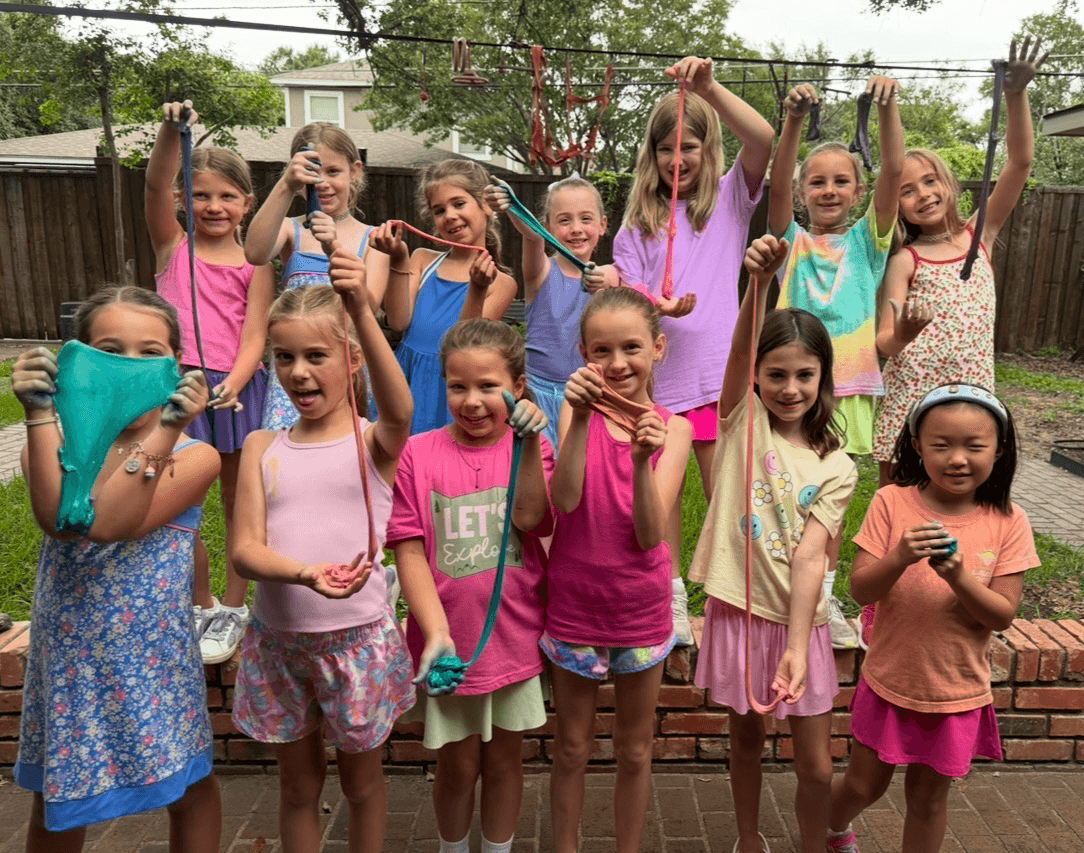 Group of children outdoors, smiling and holding a jump rope, standing on a brick patio with a wooden fence and trees behind.