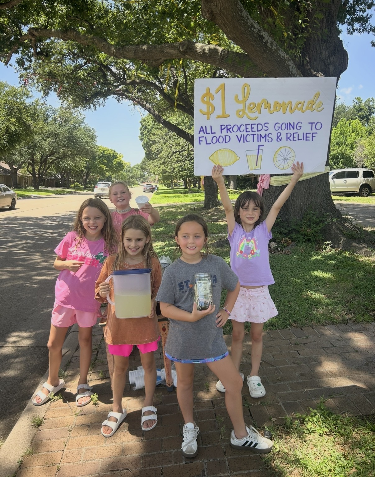 Group of six children standing outdoors under a tree, holding a sign supporting flood victims, with green grass and parked cars in background.