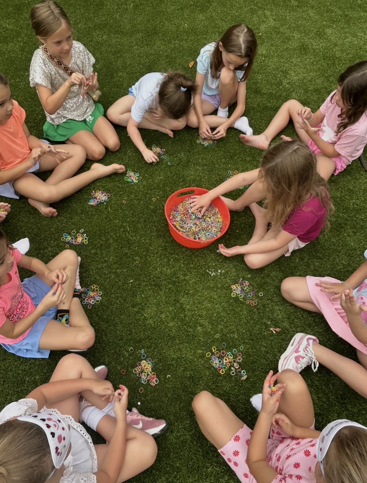 Group of children sitting on grass, playing with colorful beads and small objects, with a red container in the center.