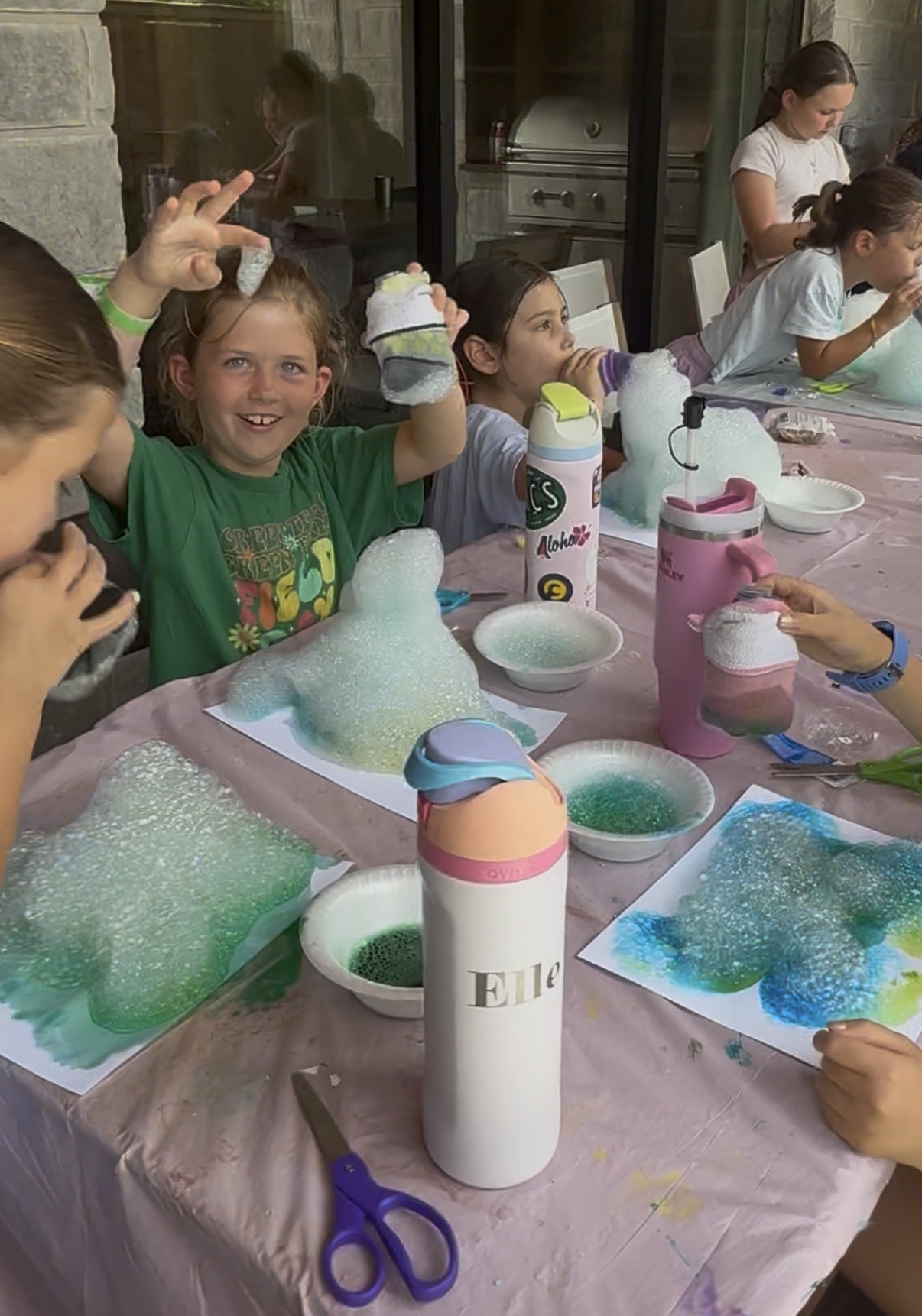 Children at a table with soap bubbles, bottles, and tools, engaging in a bubble-making activity outdoors.