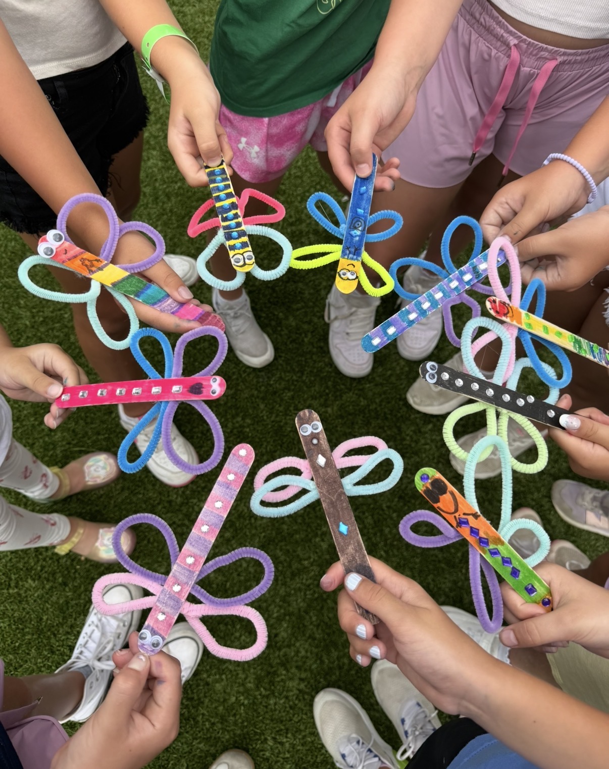 Children hold colorful butterfly-shaped hairbands with various patterns, standing in a circle outdoors on grass.