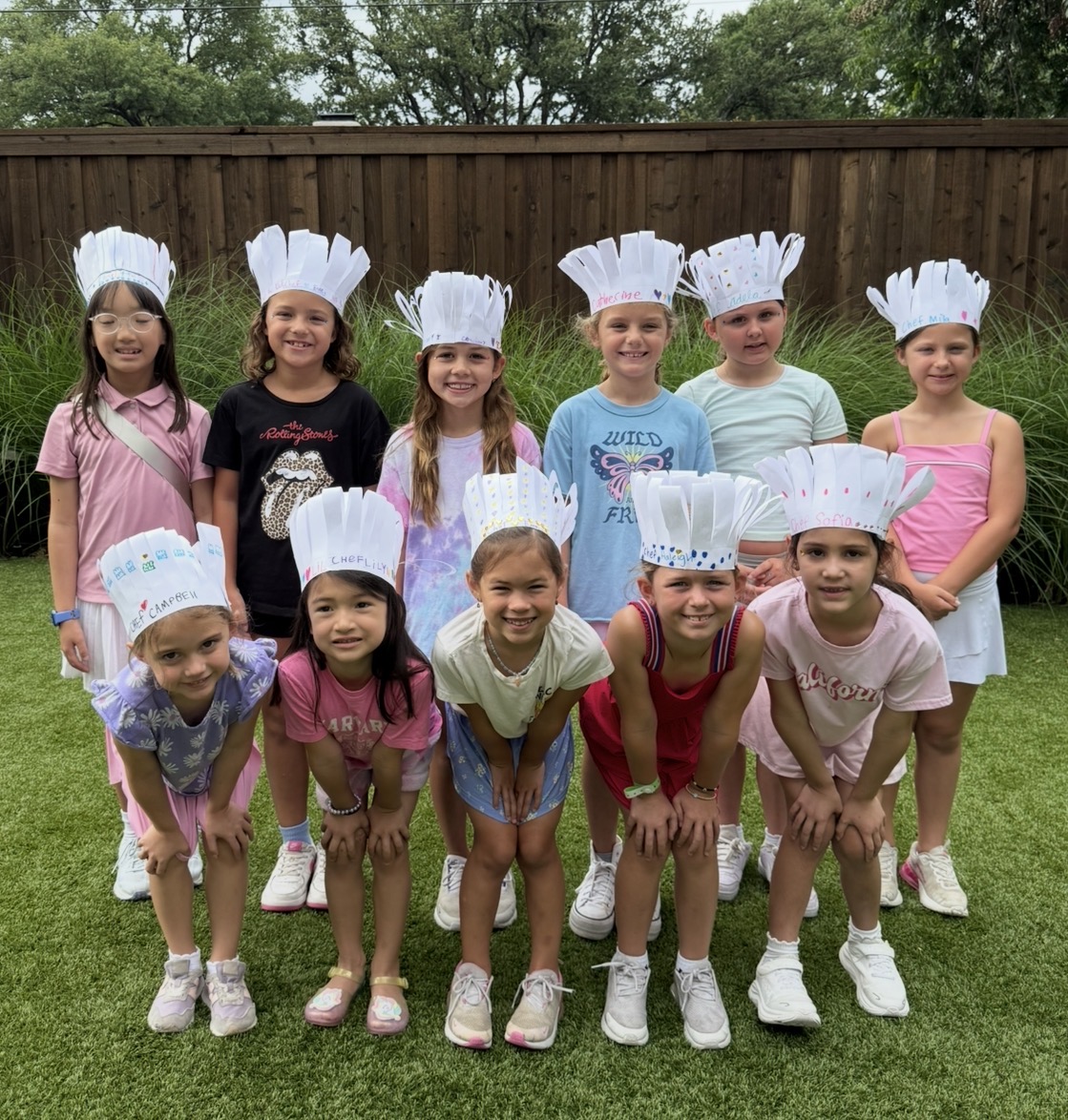 Group of ten children wearing chef hats and casual clothes, standing outdoors on grass with a wooden fence behind them.