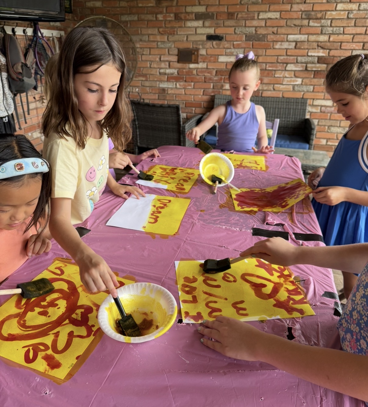 Children decorate yellow posters with red paint at a table covered with a pink tablecloth, against a brick wall background.