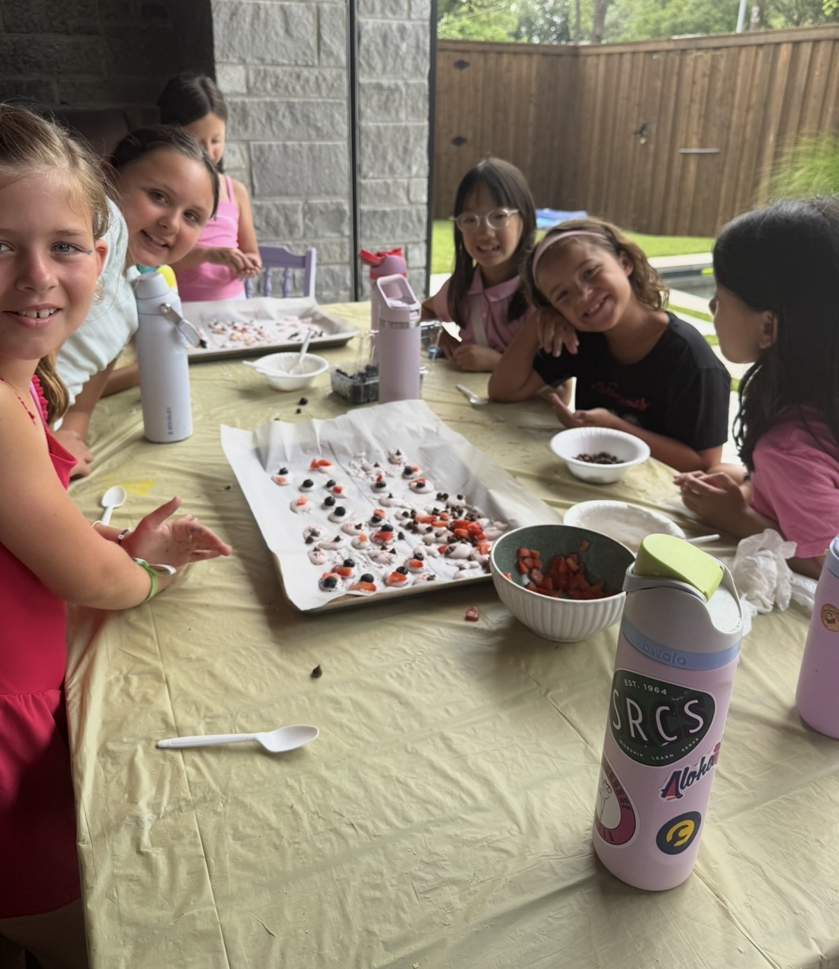 Group of children gathered around a table with snacks and drinks outdoors, smiling and enjoying a gathering.