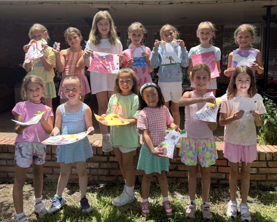 Group of children holding colorful paintings outdoors, standing on grass in front of a brick wall, smiling.