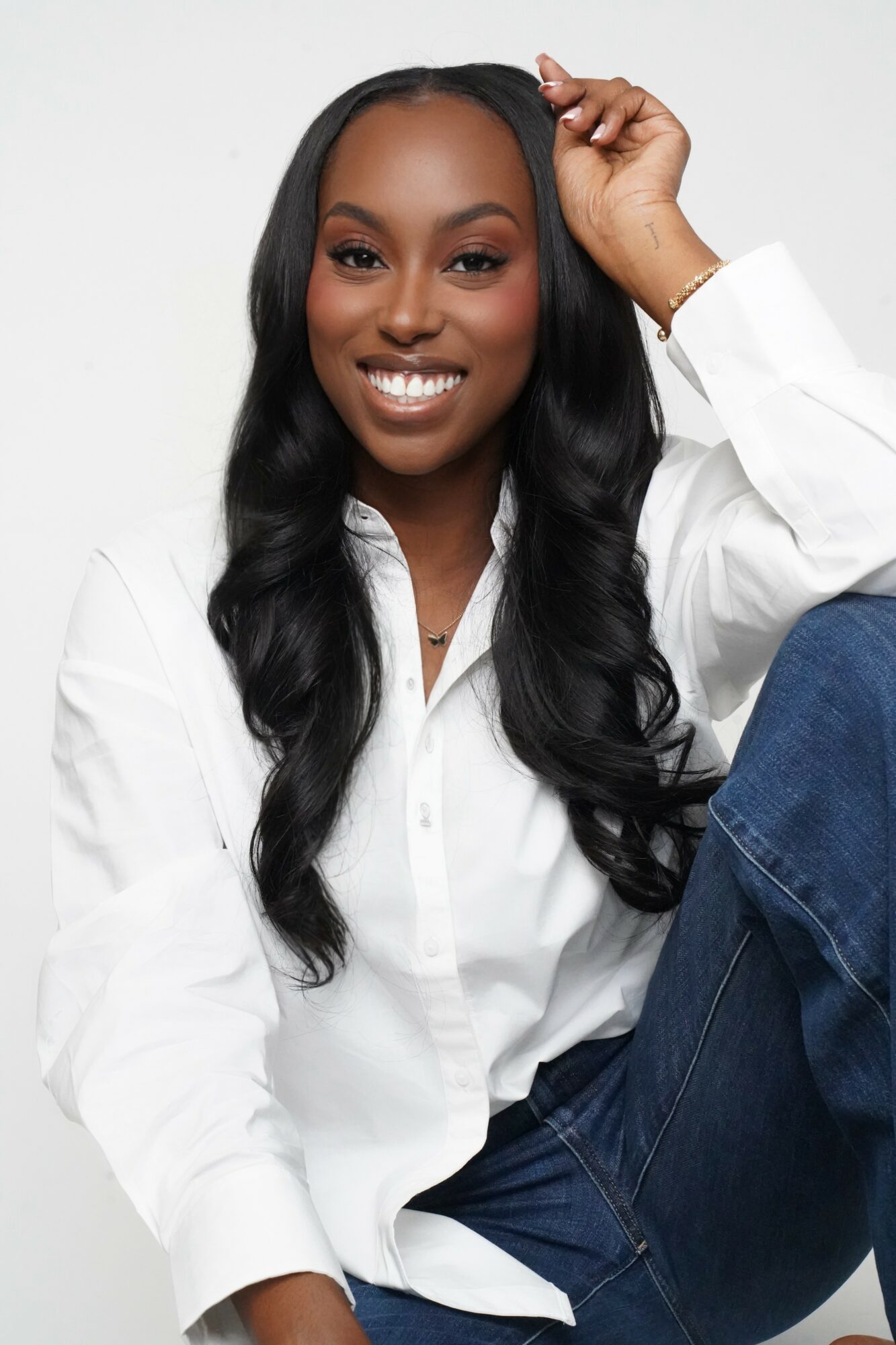 Smiling woman with long black hair, wearing a white shirt, sitting against a plain background.