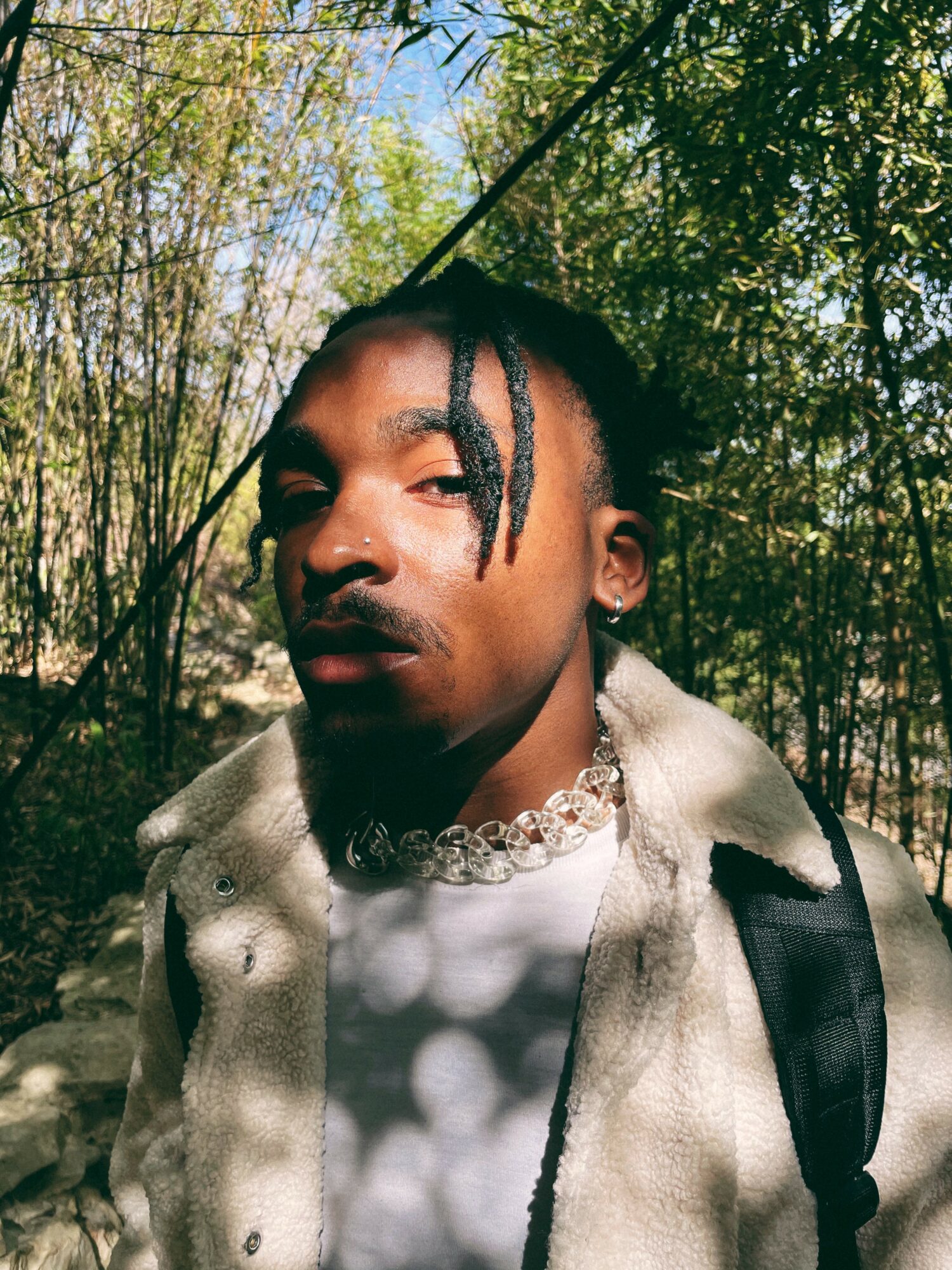 Young person with dreadlocks outdoors among trees, wearing a light-colored jacket and a choker necklace.