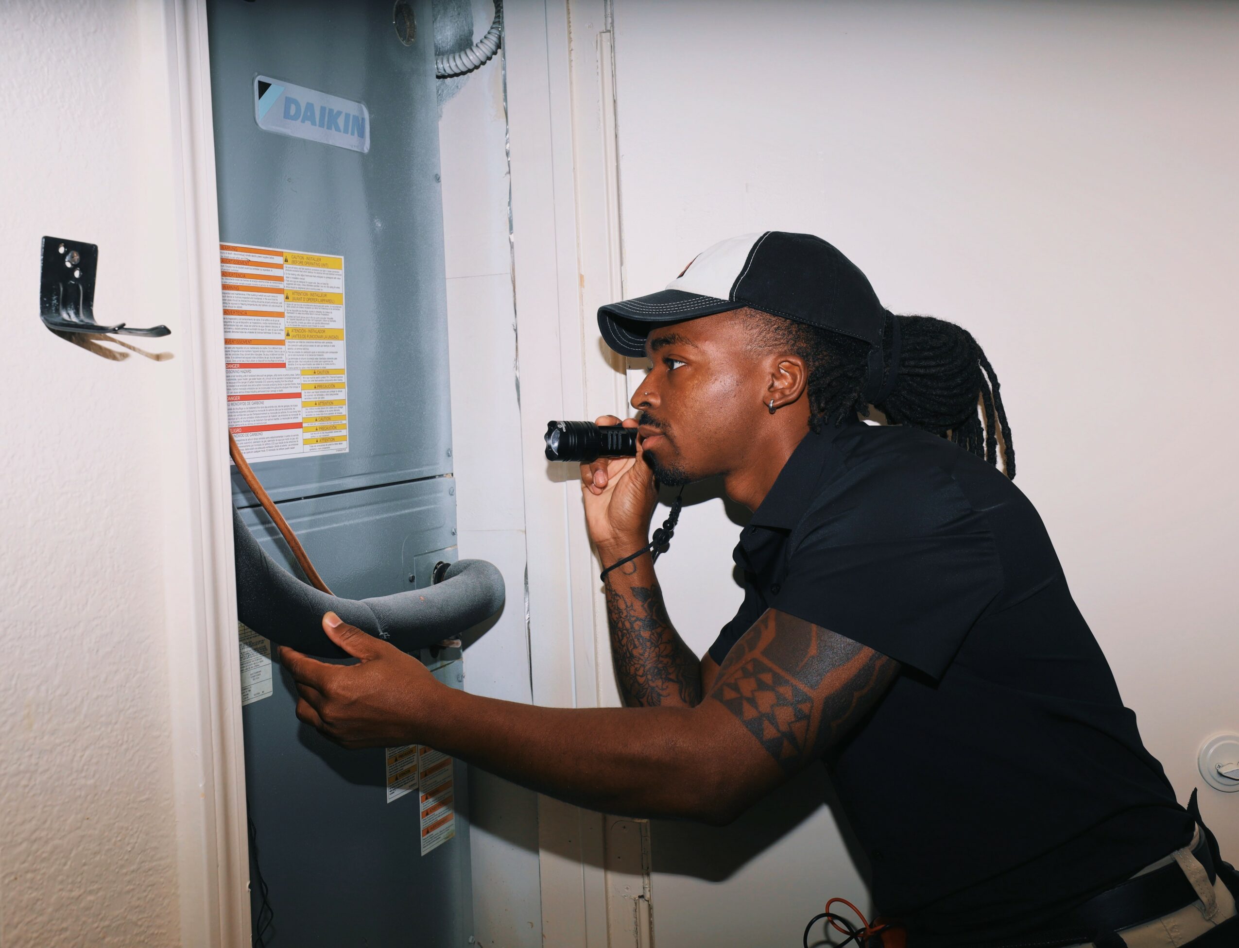 Person inspecting an electrical panel, holding a flashlight and a clipboard, wearing a black cap and shirt.