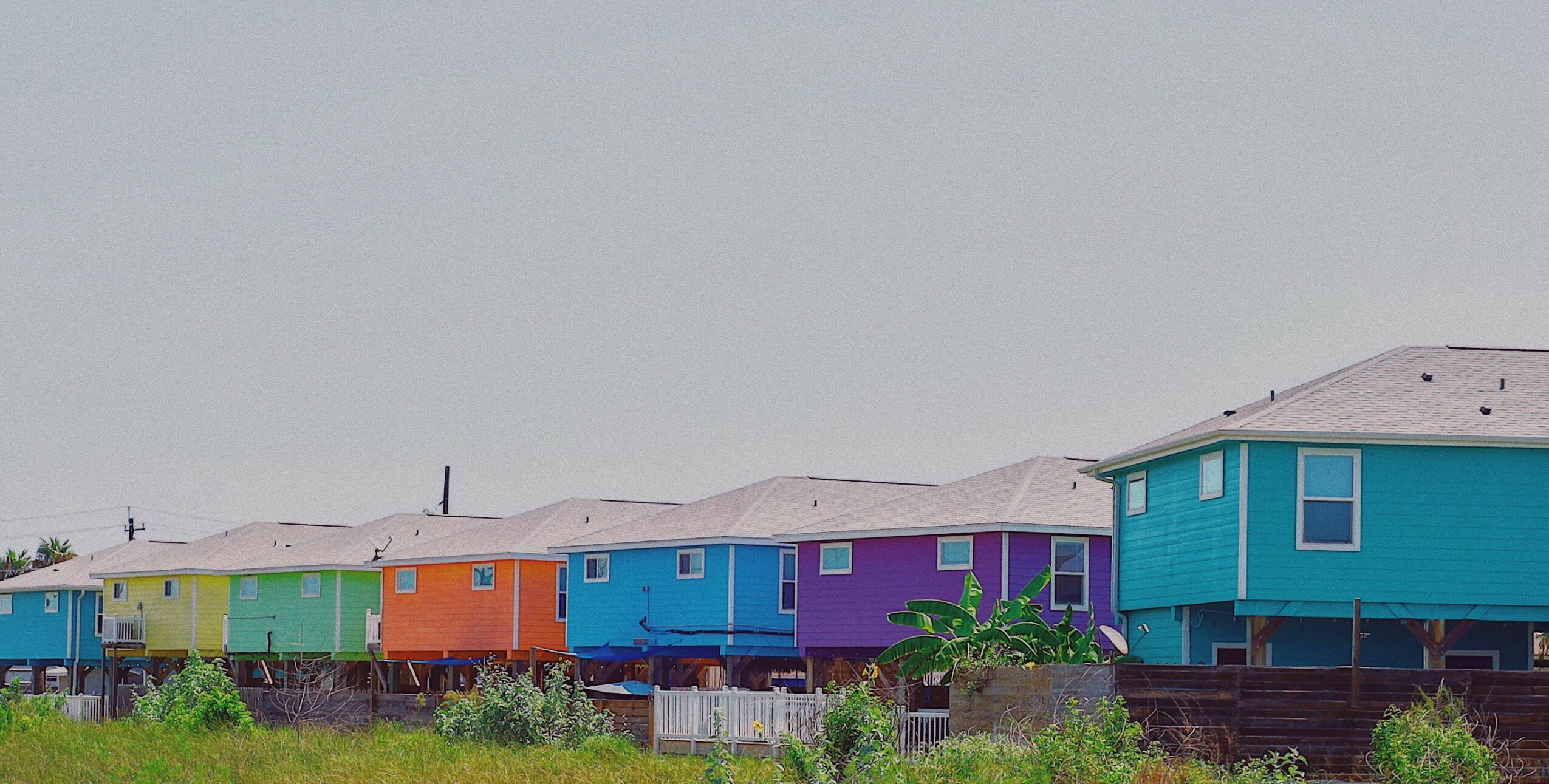 Line of colorful houses on stilts, with a grassy foreground and a plain sky background.