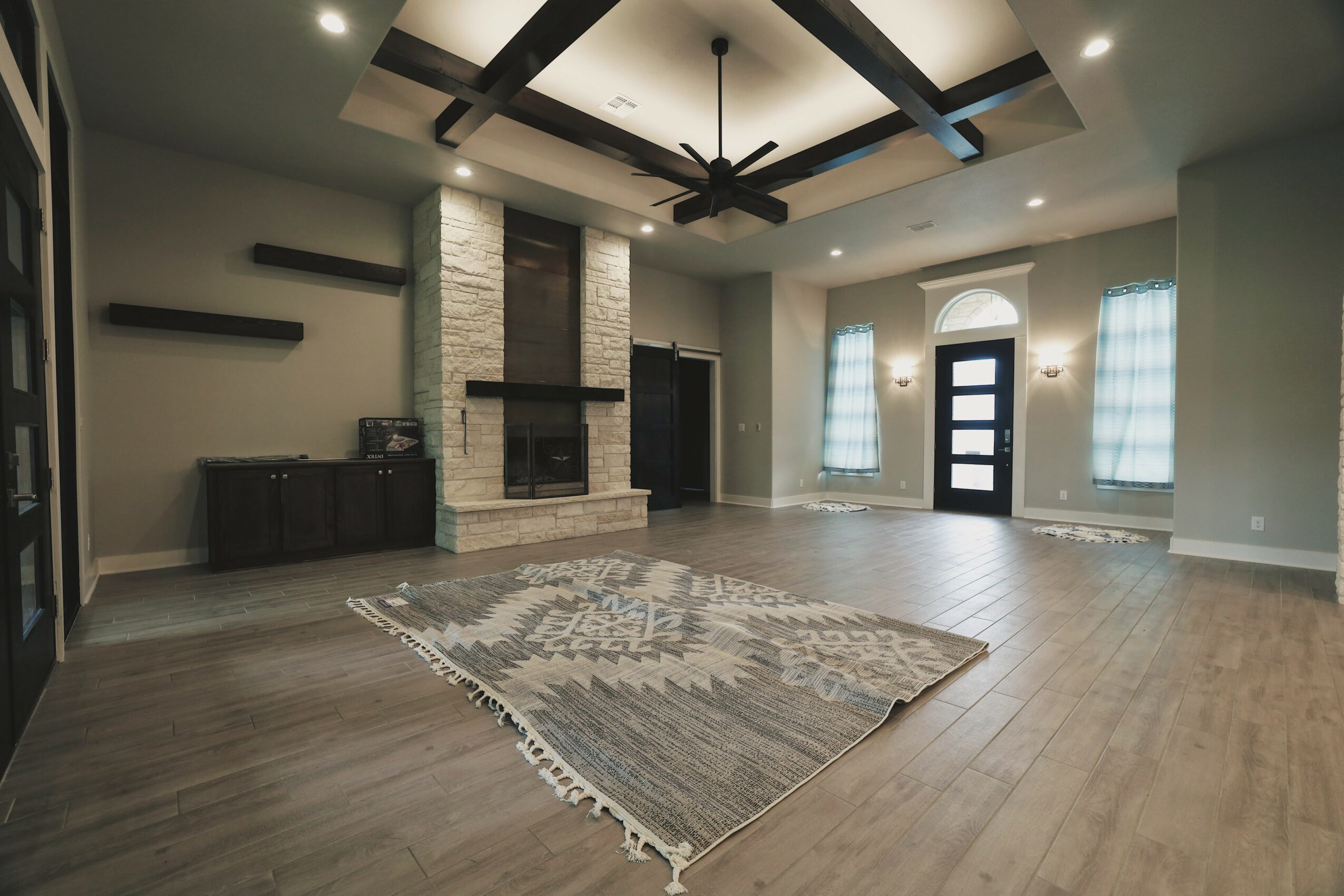 Living room with a stone fireplace, ceiling fan, large windows, and a patterned rug on hardwood floor.