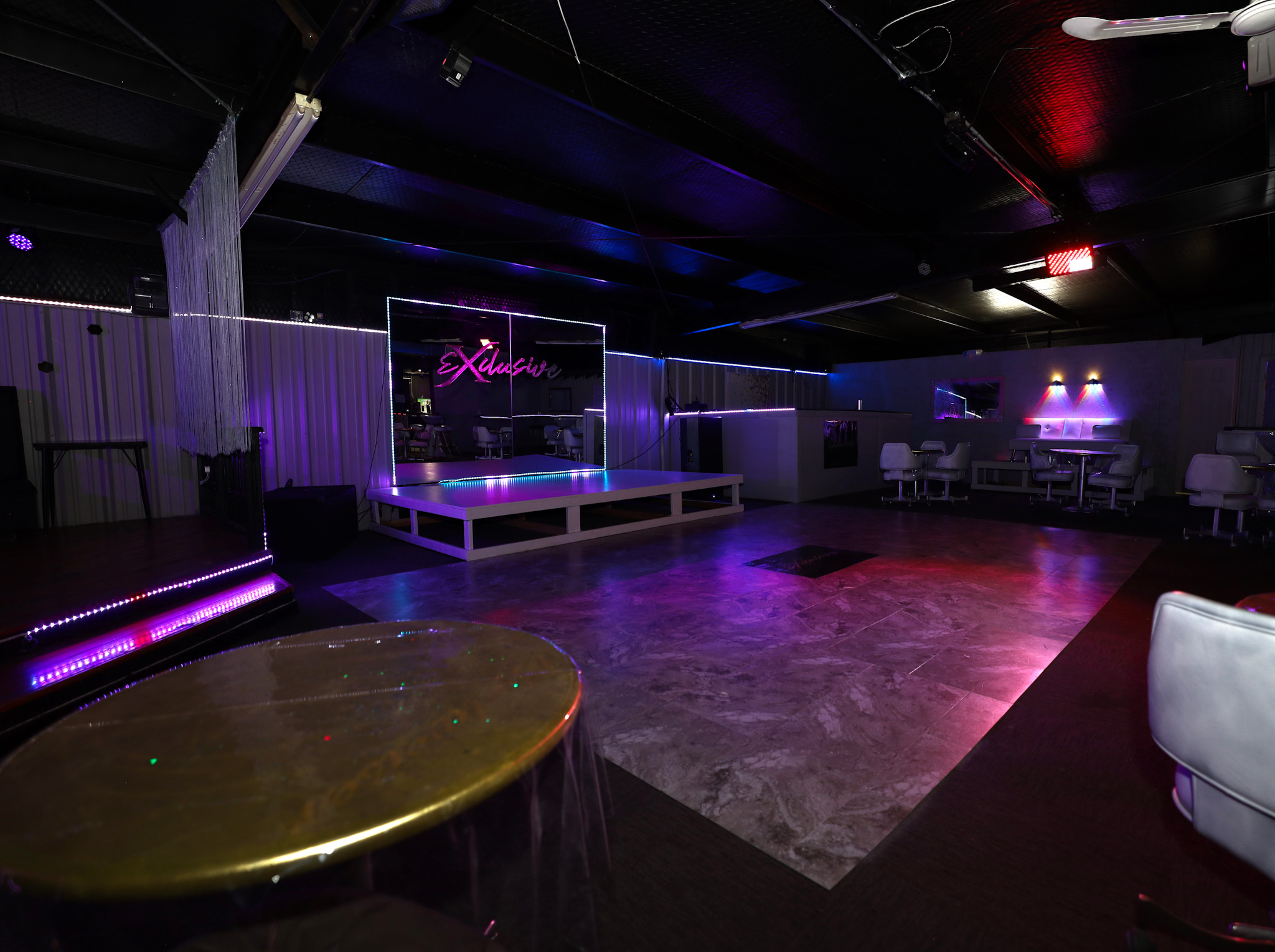 Empty nightclub stage with purple and pink lighting, surrounded by seating areas and a reflective floor.