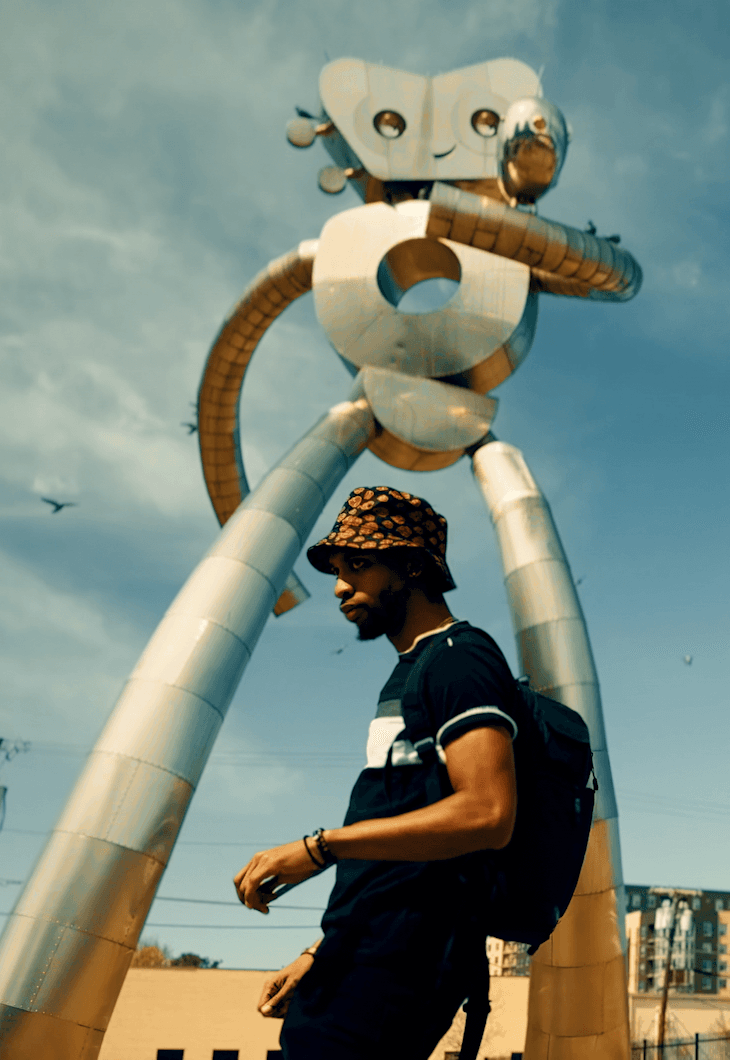 Person walking past a large robot sculpture outdoors, with a cloudy sky background.