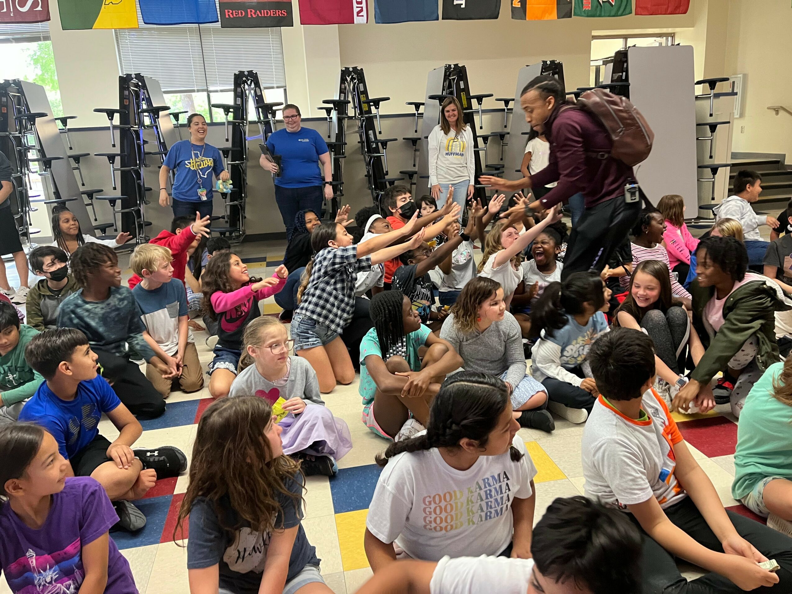 Group of children sitting on the floor in a classroom, reaching out to a person standing and handing something to a child.