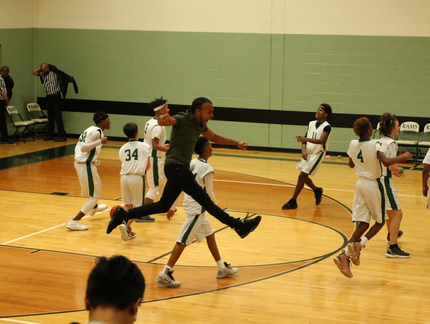 Group of children playing basketball on indoor court, some running and others standing, with a coach or adult nearby.