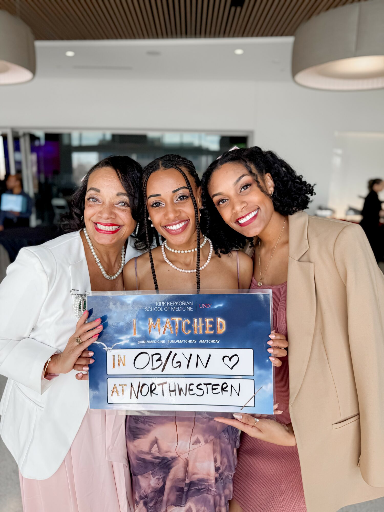 Three women smiling and holding a sign that says 'Matched in OB/GYN at Northwestern' in a modern indoor setting.