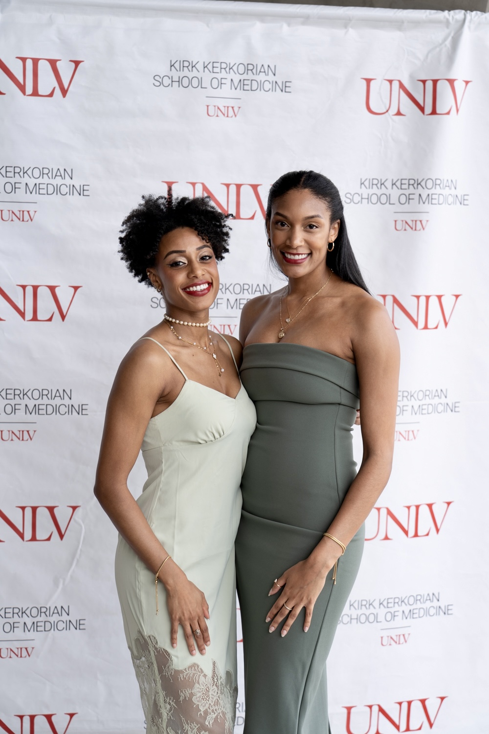 Two women in formal dresses standing in front of a UNLV backdrop, smiling, one with curly hair and the other with straight hair.