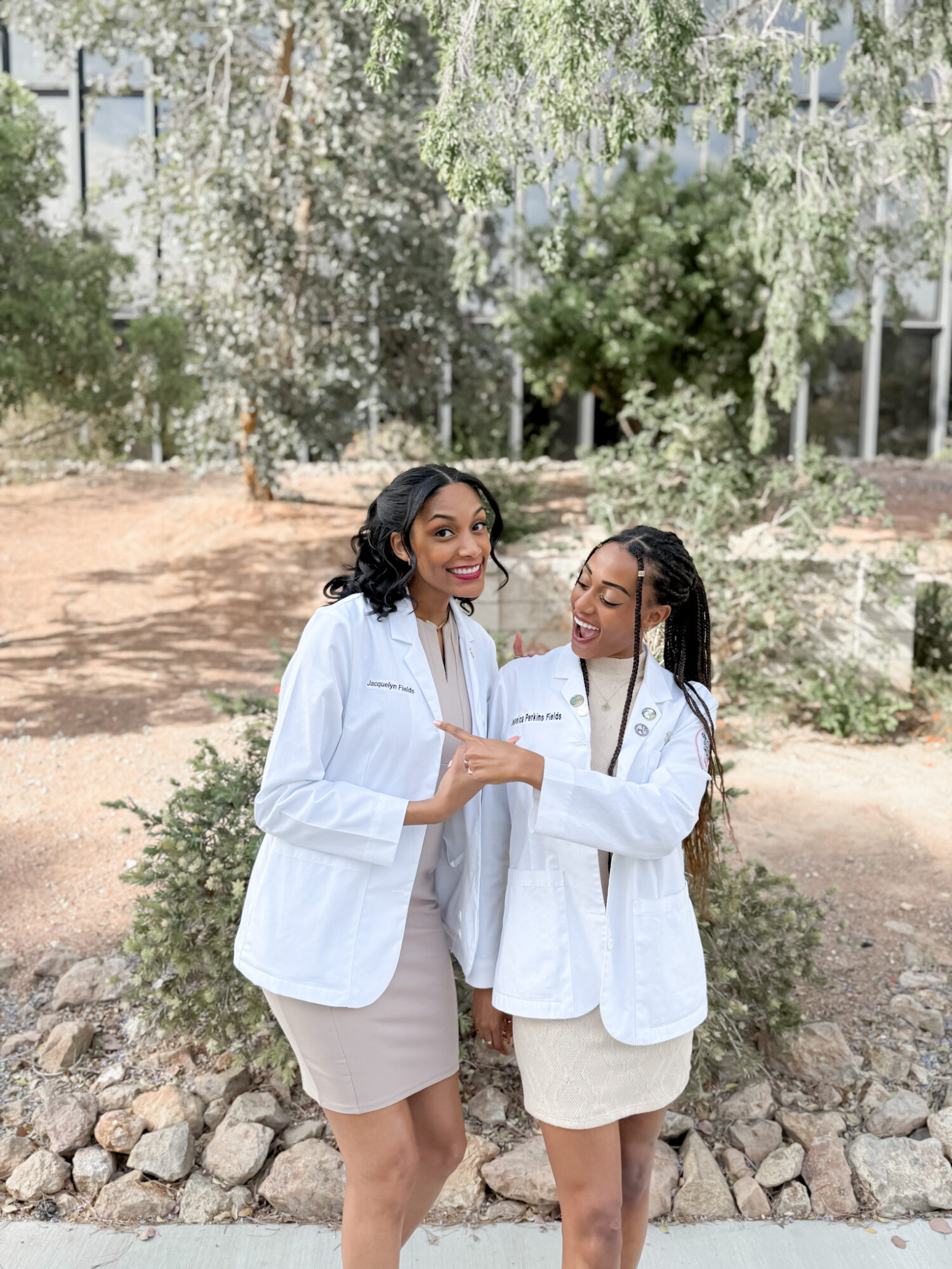 Two women stand outdoors in front of trees and bushes, smiling and talking, wearing white blazers and beige skirts.