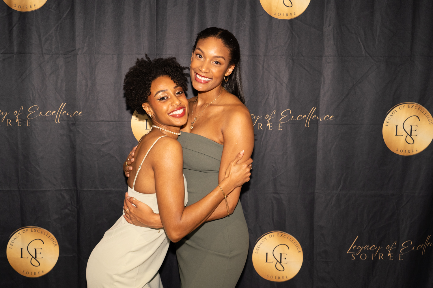 Two women smiling and hugging in front of a black backdrop with gold logos, one with curly hair and the other with straight hair.