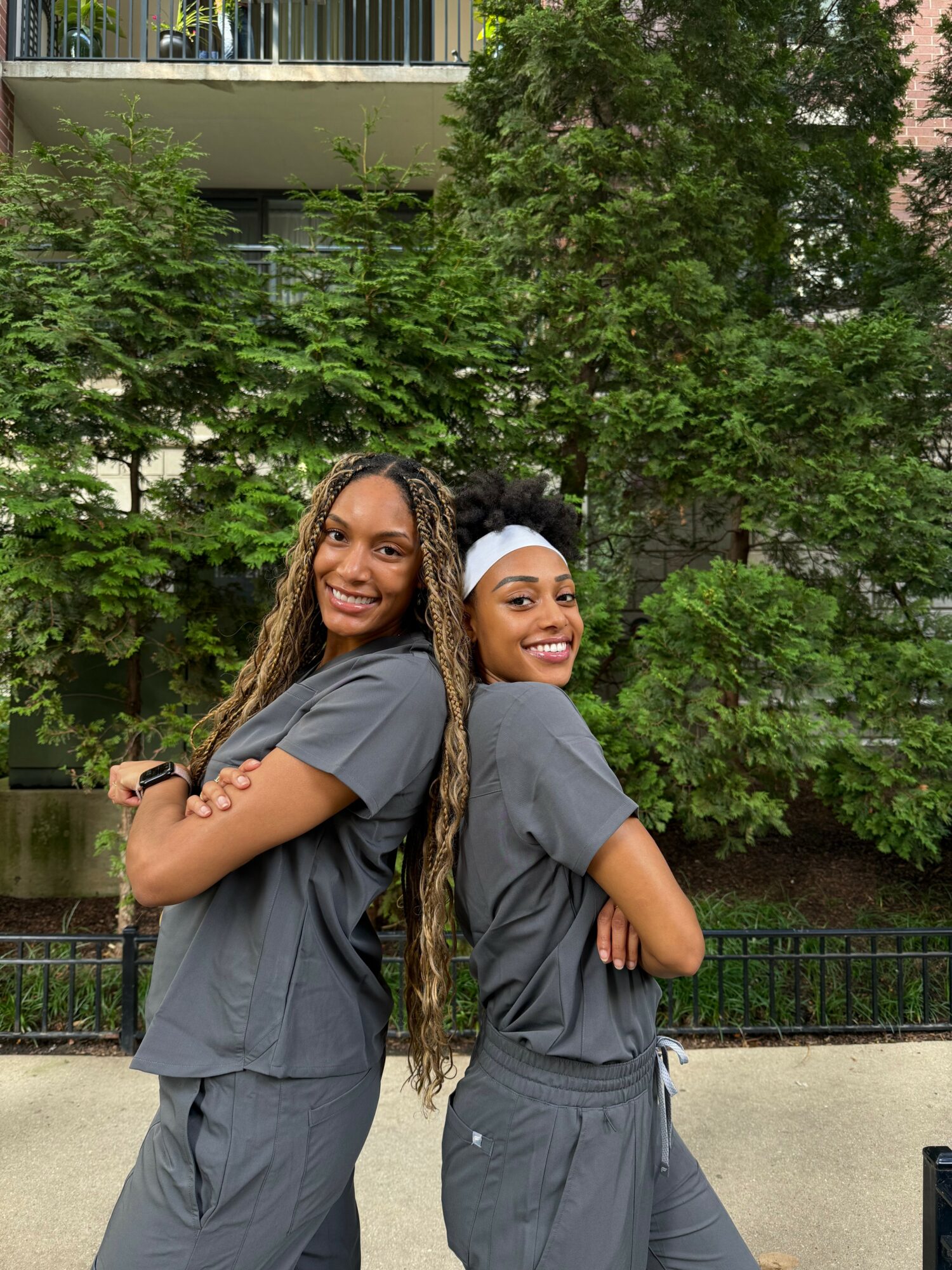 Two smiling women with long hair in gray outfits standing outdoors with trees and buildings behind them.