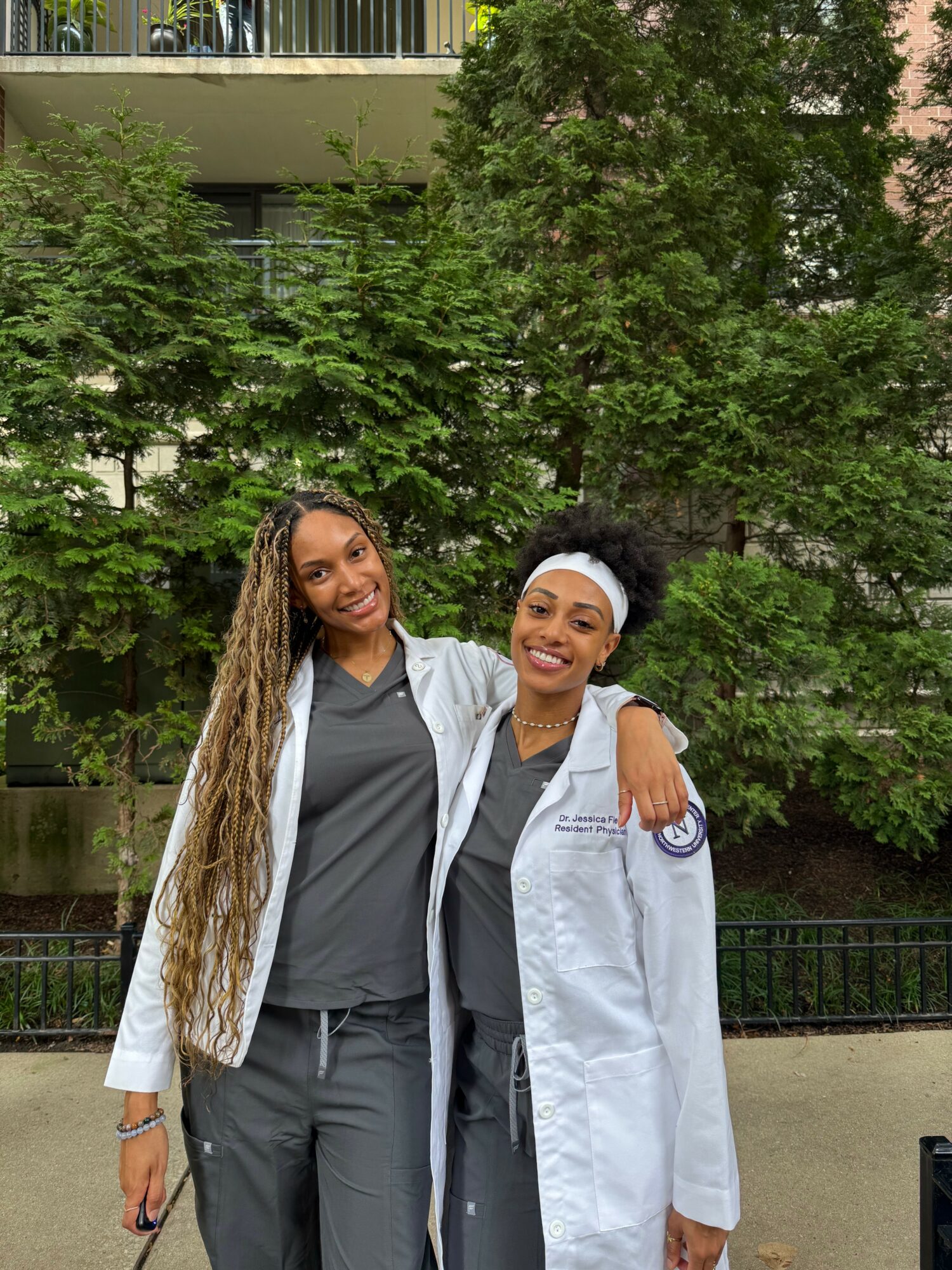 Two women smiling outdoors, one with long curly hair and the other in a white coat, standing close together with trees in background.