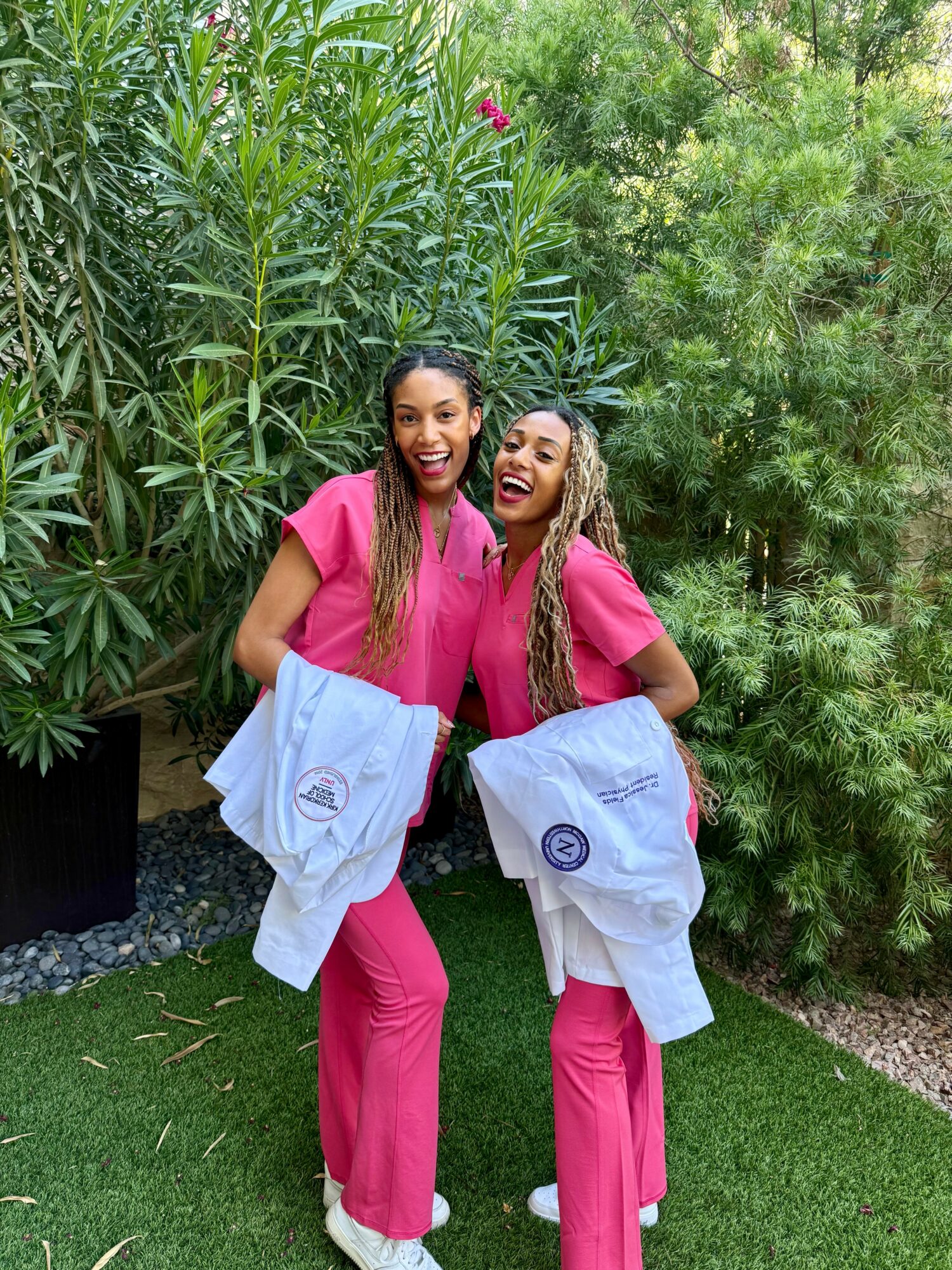 Two women in pink scrubs smiling outdoors, holding white jackets, standing on grass with green plants in background.