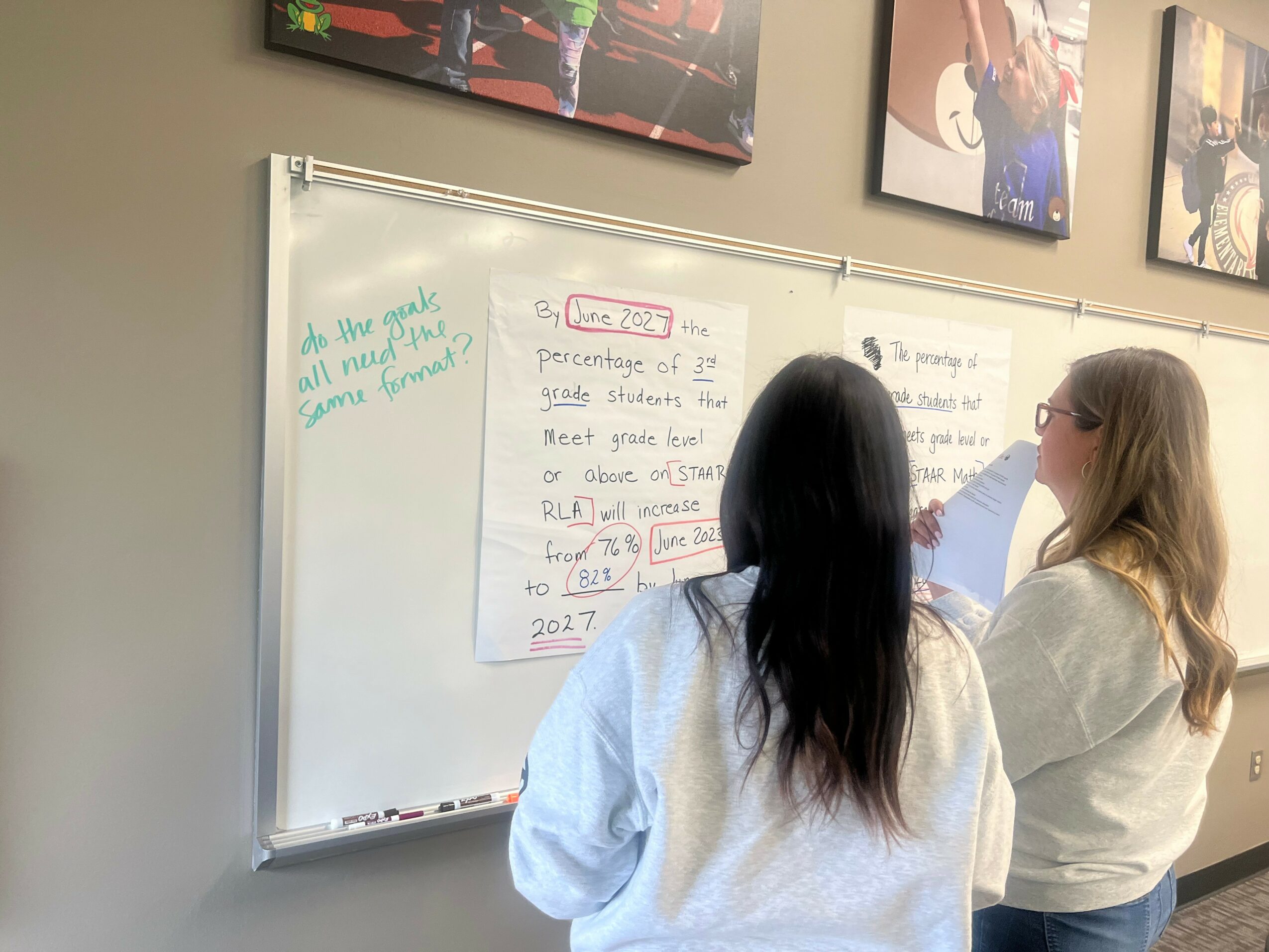 Two women stand in front of a whiteboard with handwritten notes, one pointing at the board, in a classroom setting.