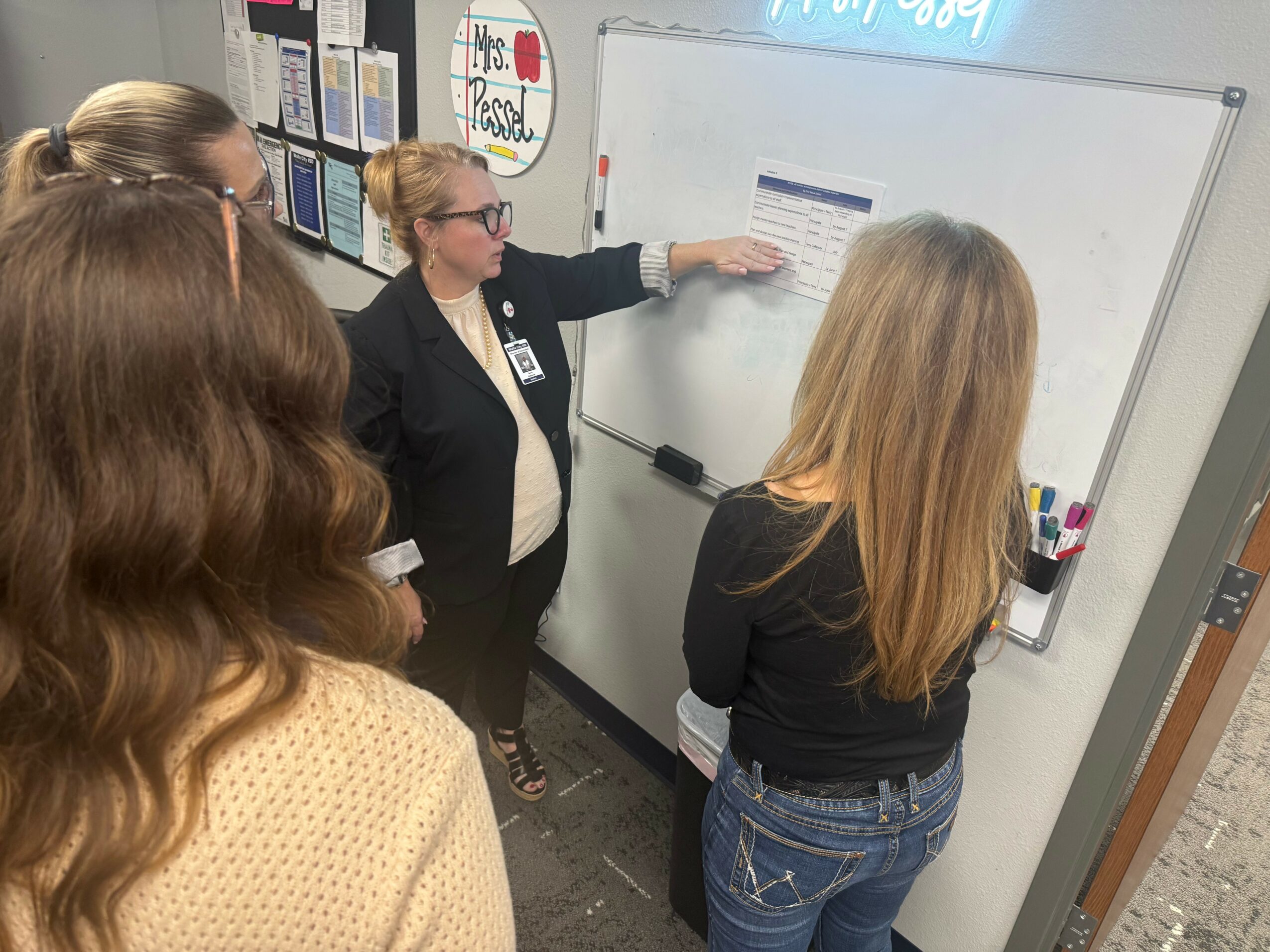 Four women in a classroom, one pointing at a whiteboard with papers, others listening attentively.