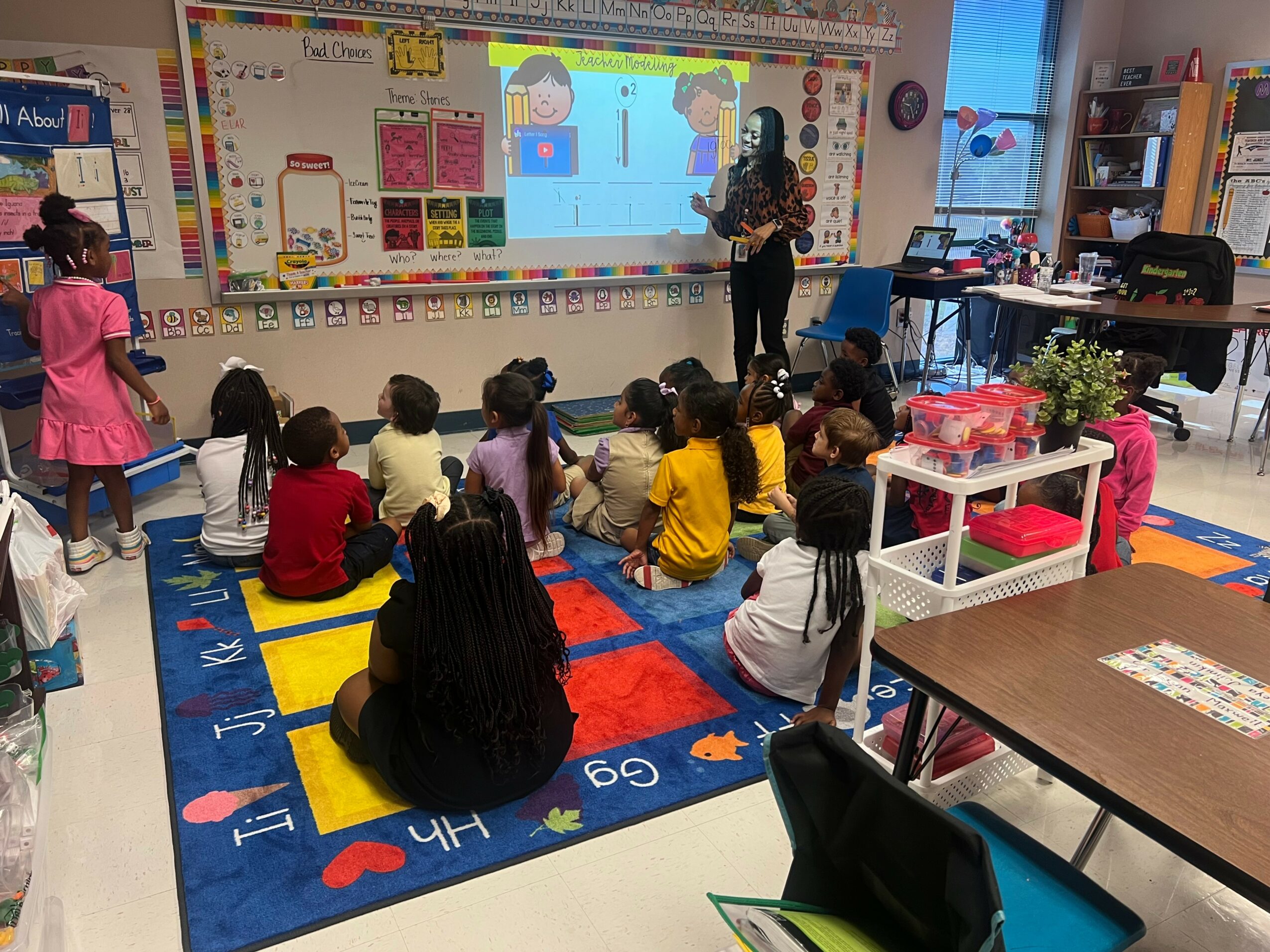 Teacher standing in front of a whiteboard with children sitting on a colorful rug, classroom with bookshelves and desks.