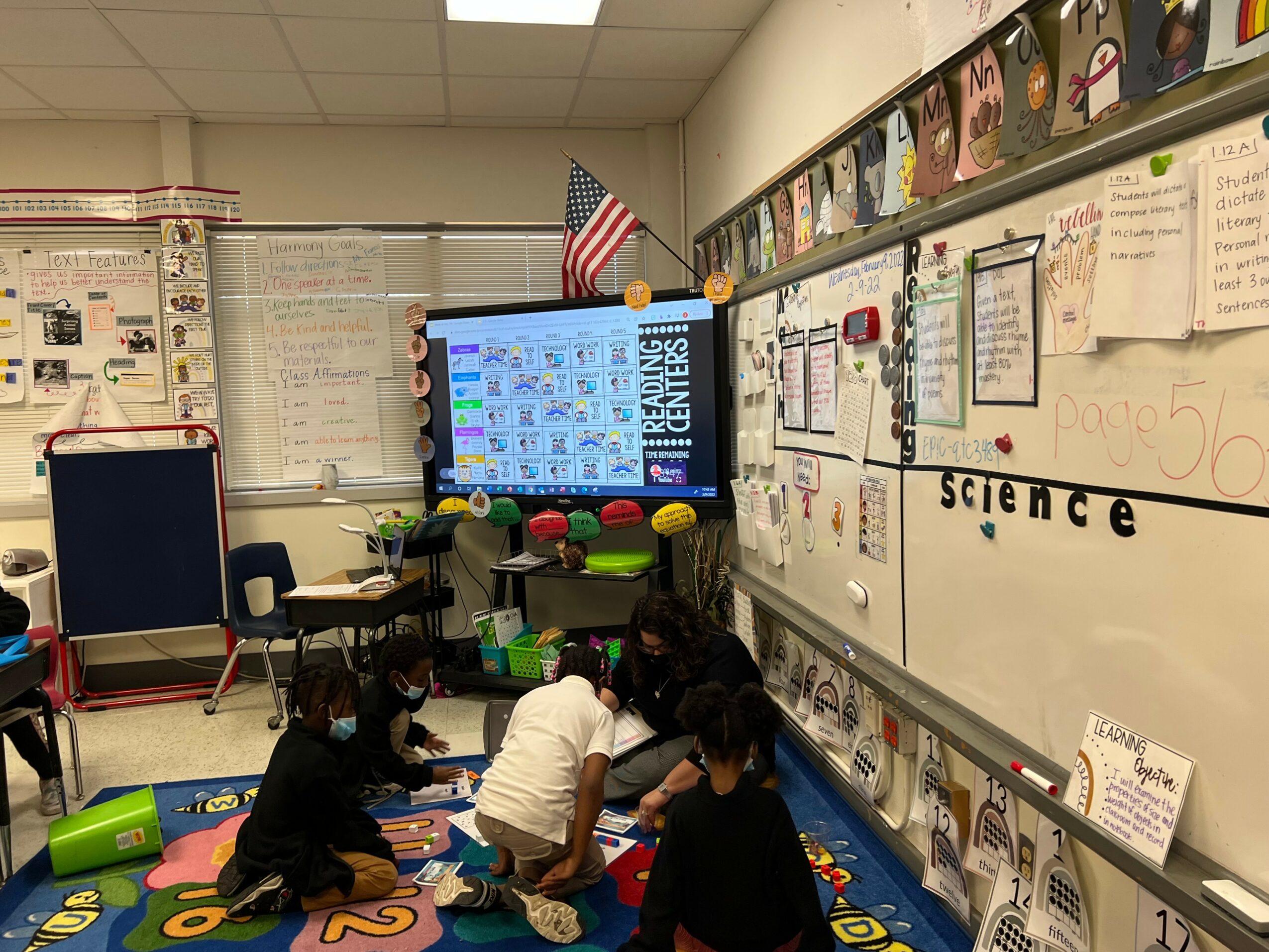 Children gather on a colorful rug in a classroom, working on a science activity near a whiteboard and a digital screen.