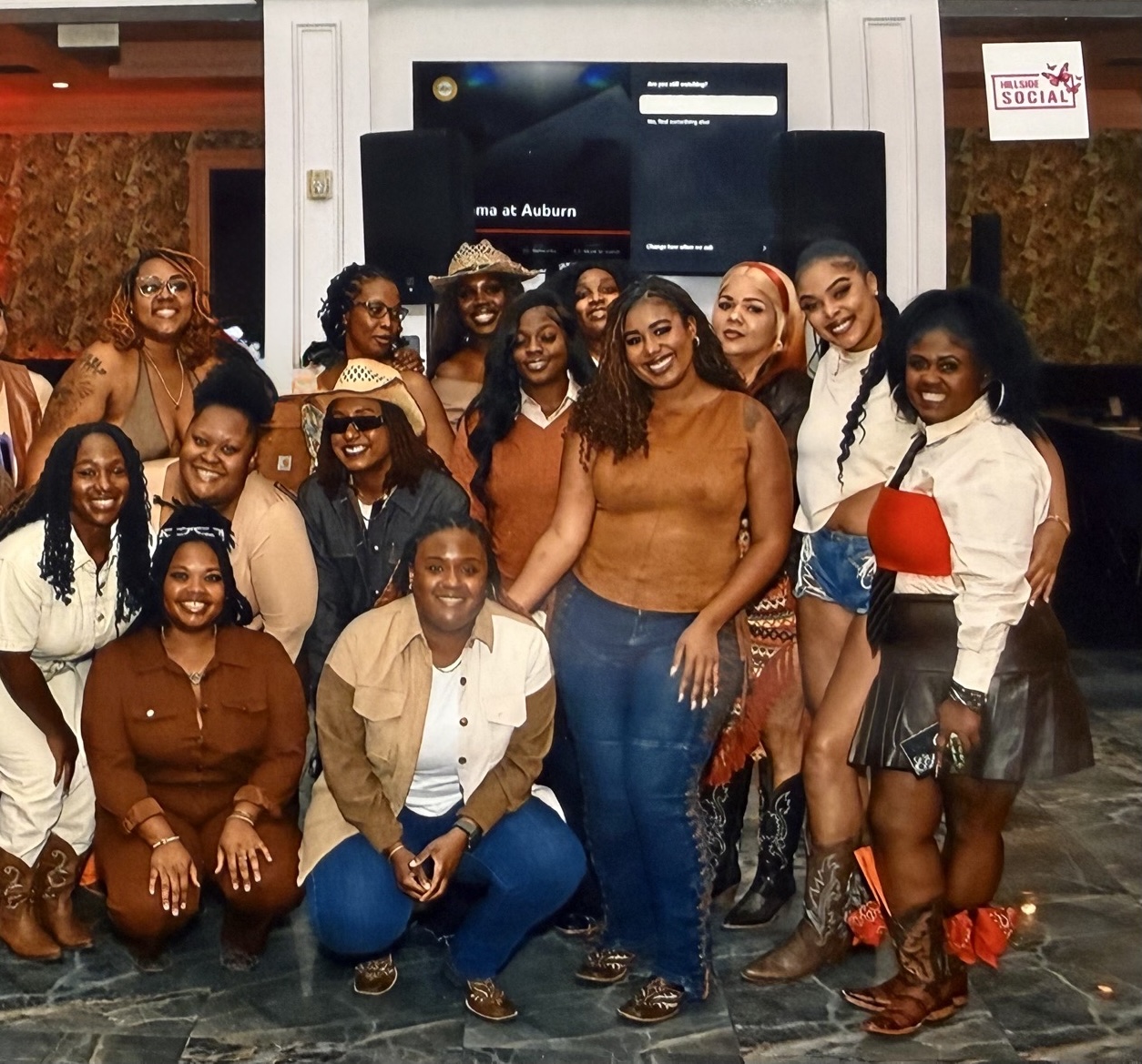 Group of women and girls posing indoors in front of a screen, smiling, with diverse clothing styles and accessories.