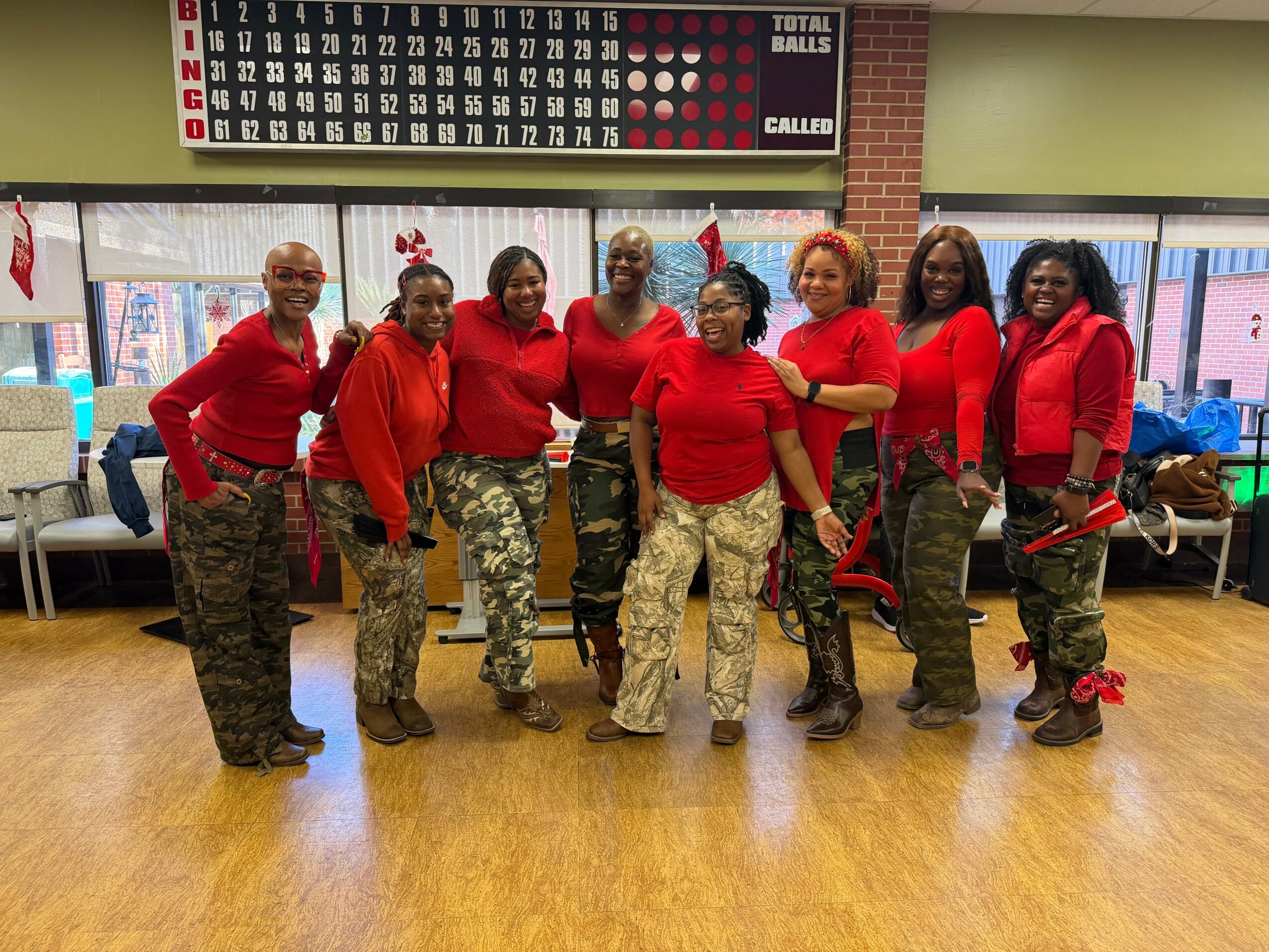 Group of women in red tops and camouflage pants standing indoors, smiling, with holiday decorations and a bingo board in background.
