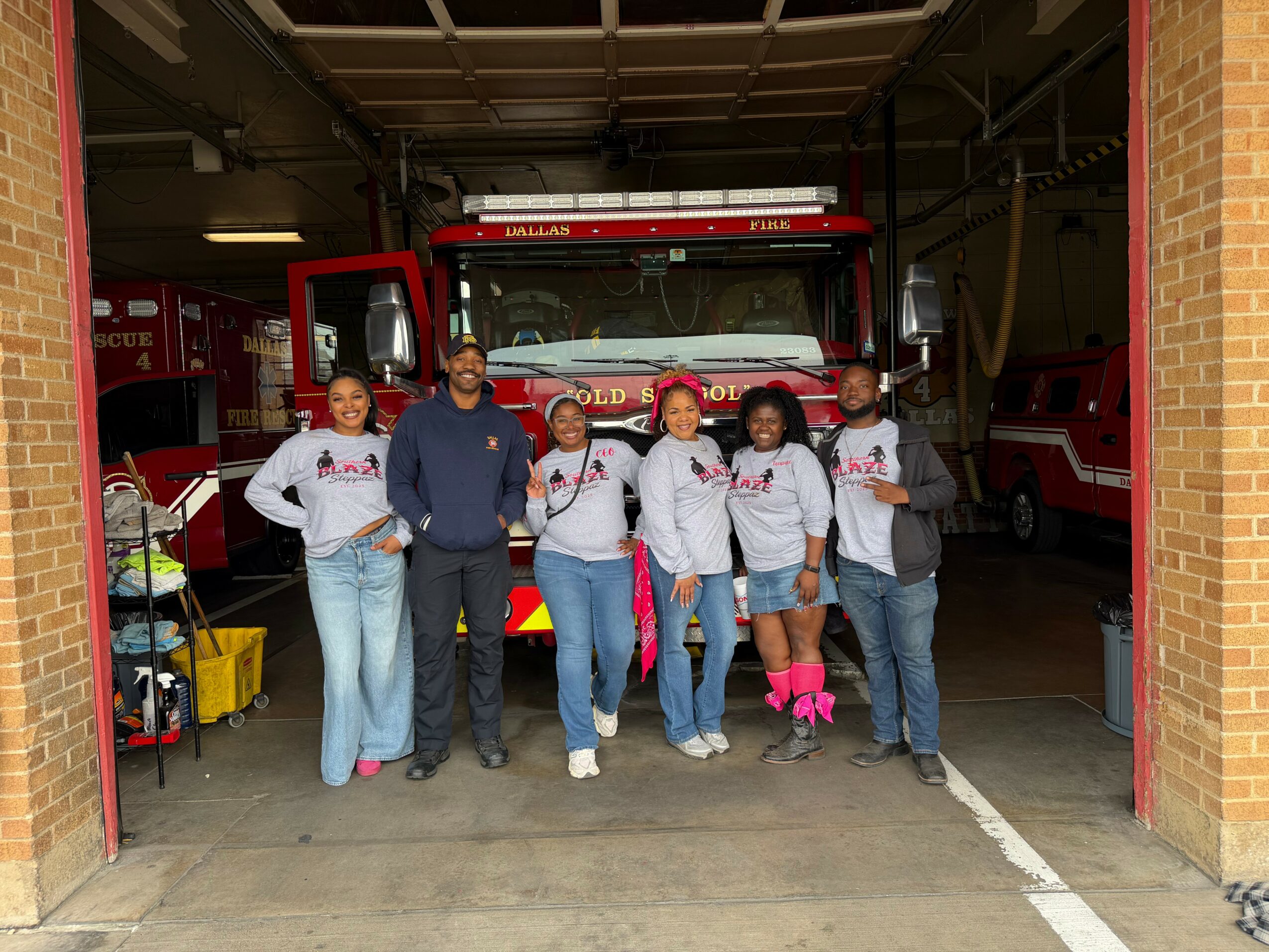 Group of six people standing in front of a fire truck inside a fire station garage, smiling and posing for the photo.