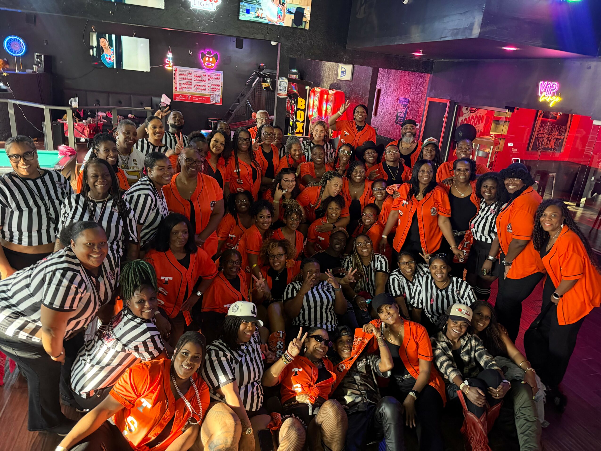 Group of people in a lively indoor setting, some wearing orange shirts and others in black and white striped shirts, with colorful lights and screens in the background.