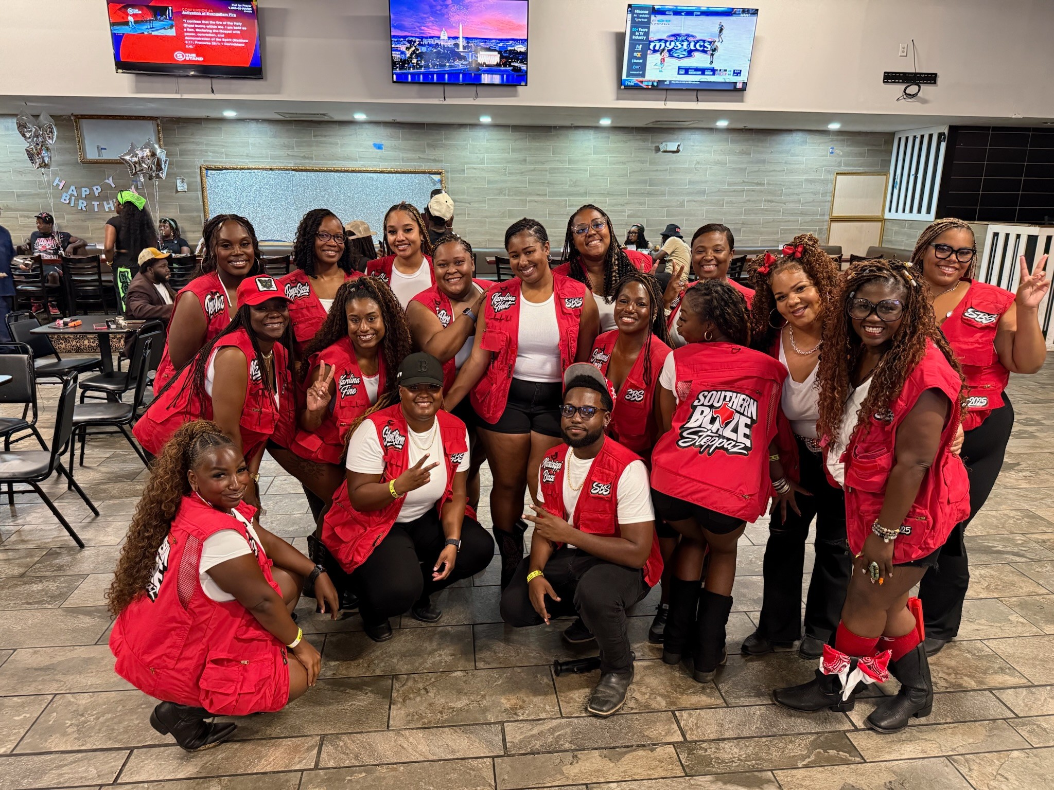 Group of young women and girls in red and white outfits posing together indoors, some making peace signs, smiling.