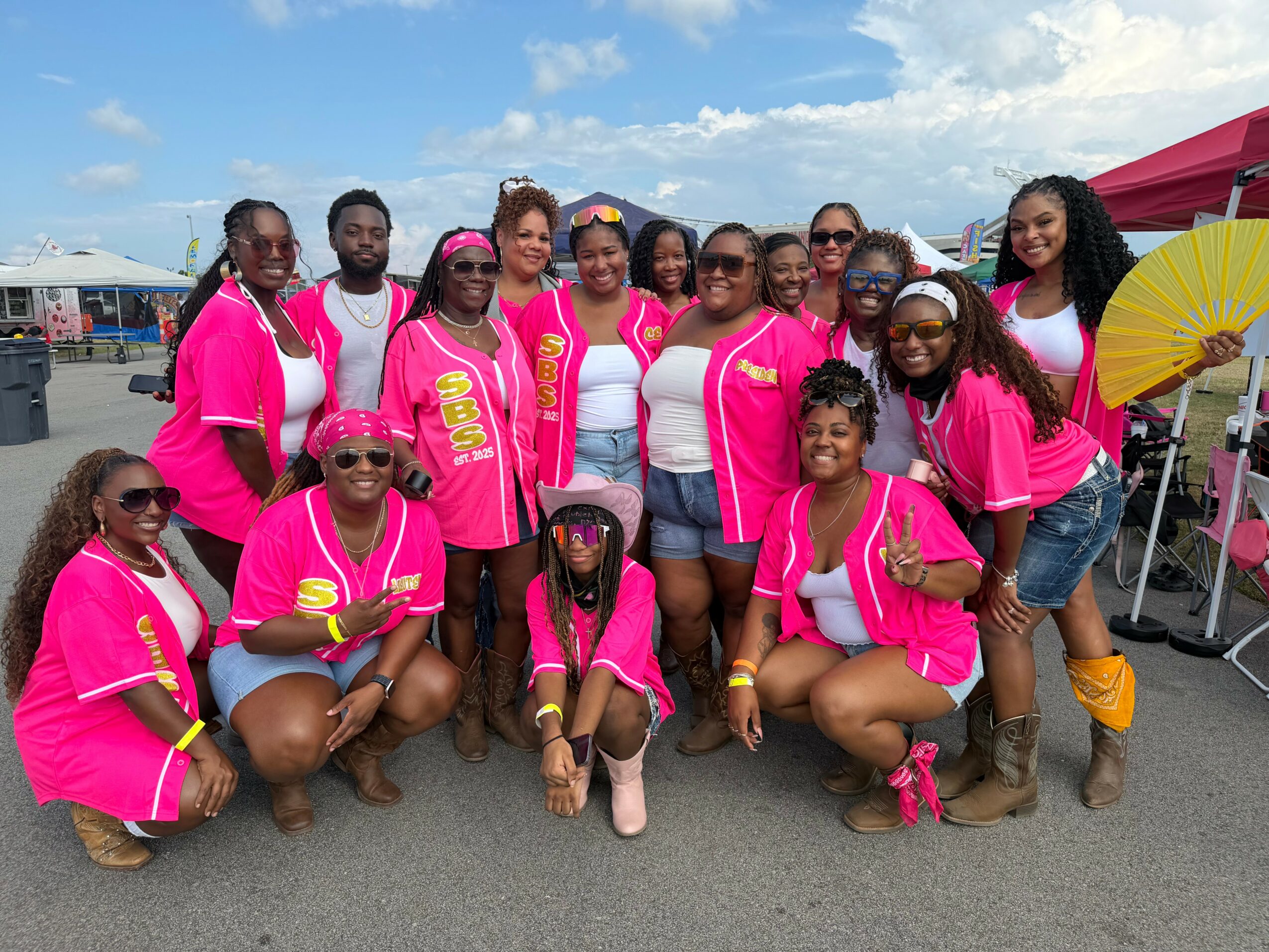 Group of people wearing pink outfits outdoors, some holding umbrellas, with tents and blue sky in background.