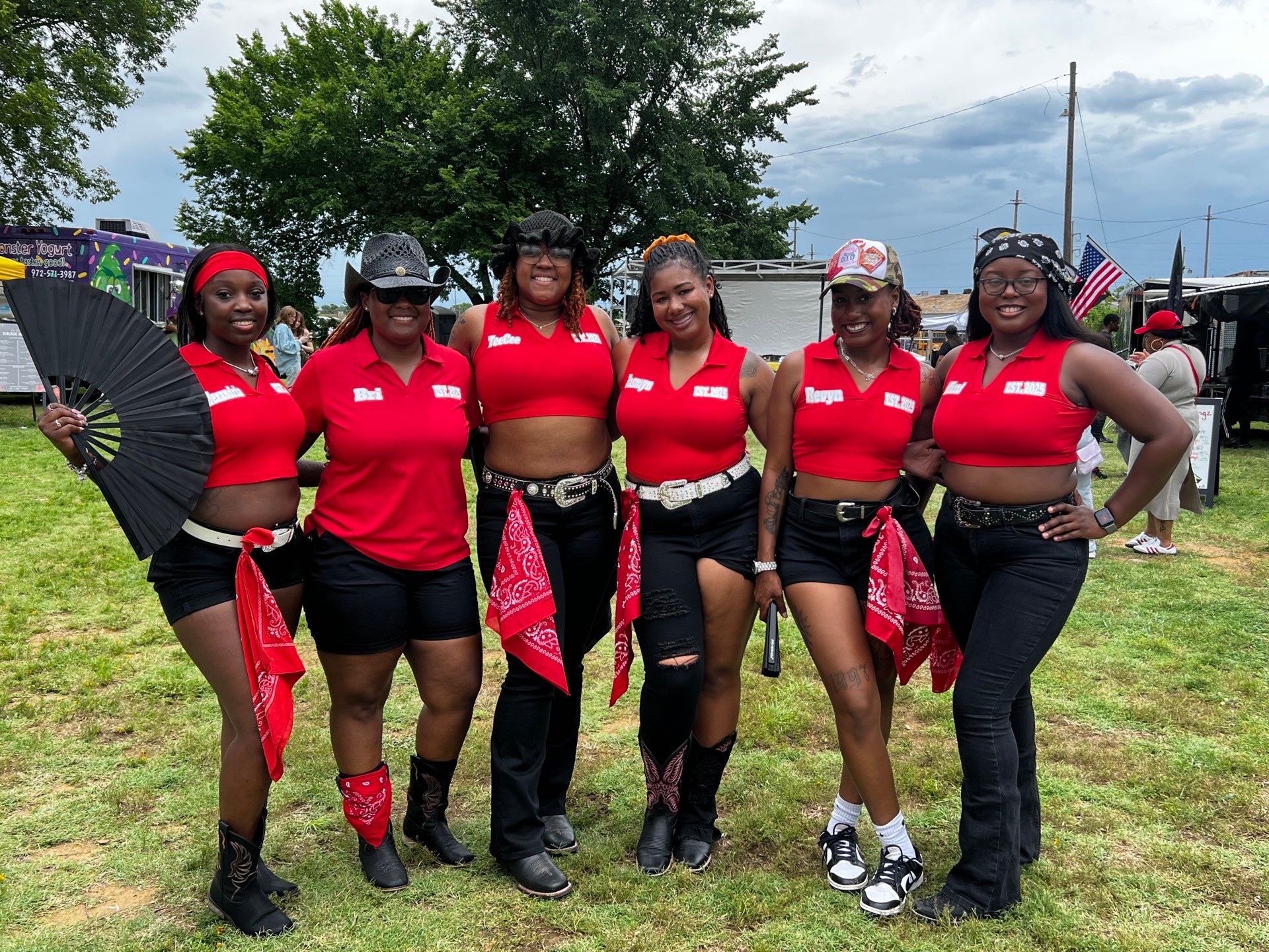 Six women in red shirts and black shorts standing outdoors, some holding fans and wearing bandanas, with trees and tents in background.