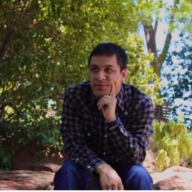 Young man sitting outdoors with trees and sunlight in background, resting chin on hand, looking thoughtful.