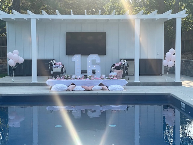 Outdoor pool with a decorated stage featuring a large number 16, balloons, and chairs, under a white pergola, with trees in background.