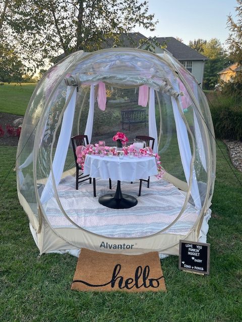 A decorated outdoor table inside a transparent dome with pink accents, set on grass with a welcome mat nearby.