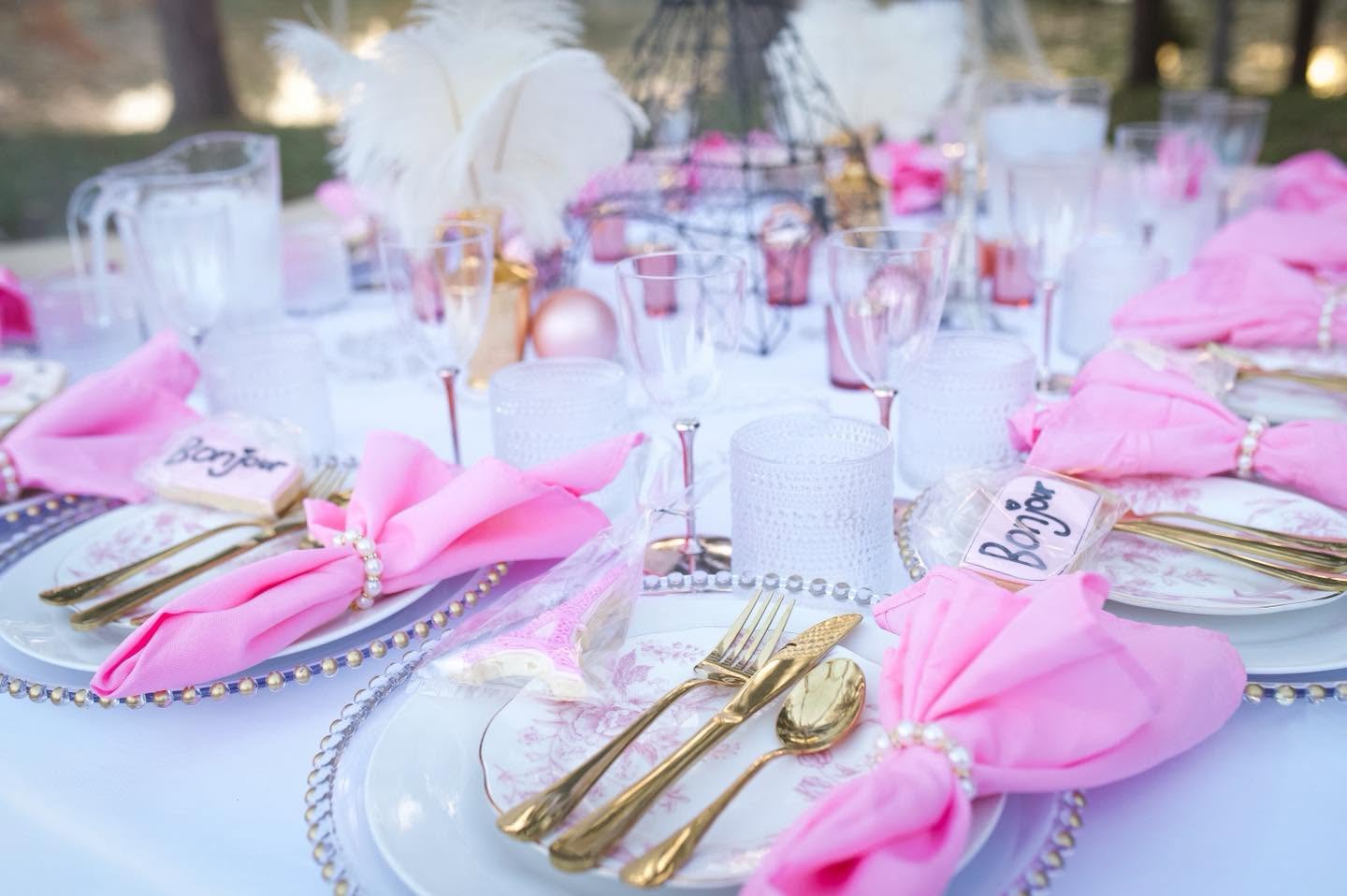 Table set with pink napkins, gold utensils, clear glasses, and decorative centerpieces, outdoors with natural light.