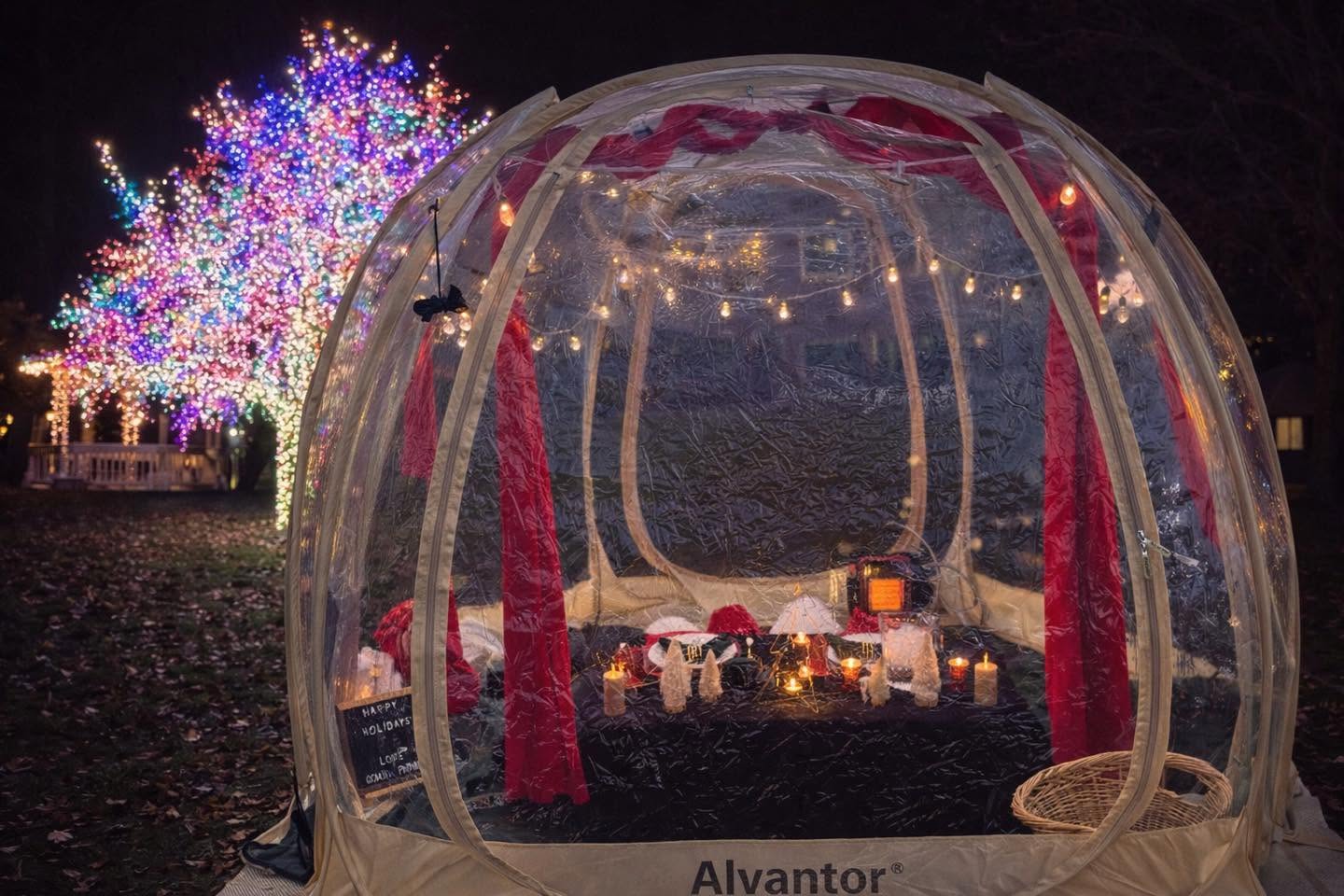 Glass igloo with red curtains and candles, outdoor decorated tree with colorful lights in background.