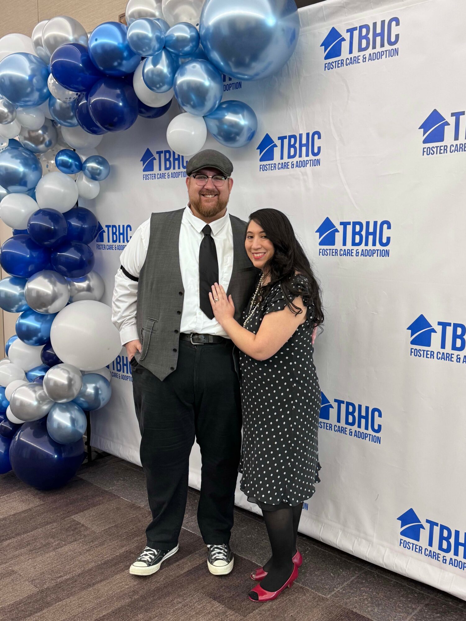 Two people standing in front of a backdrop with balloons and TBHC logo, smiling, one with arm around the other.