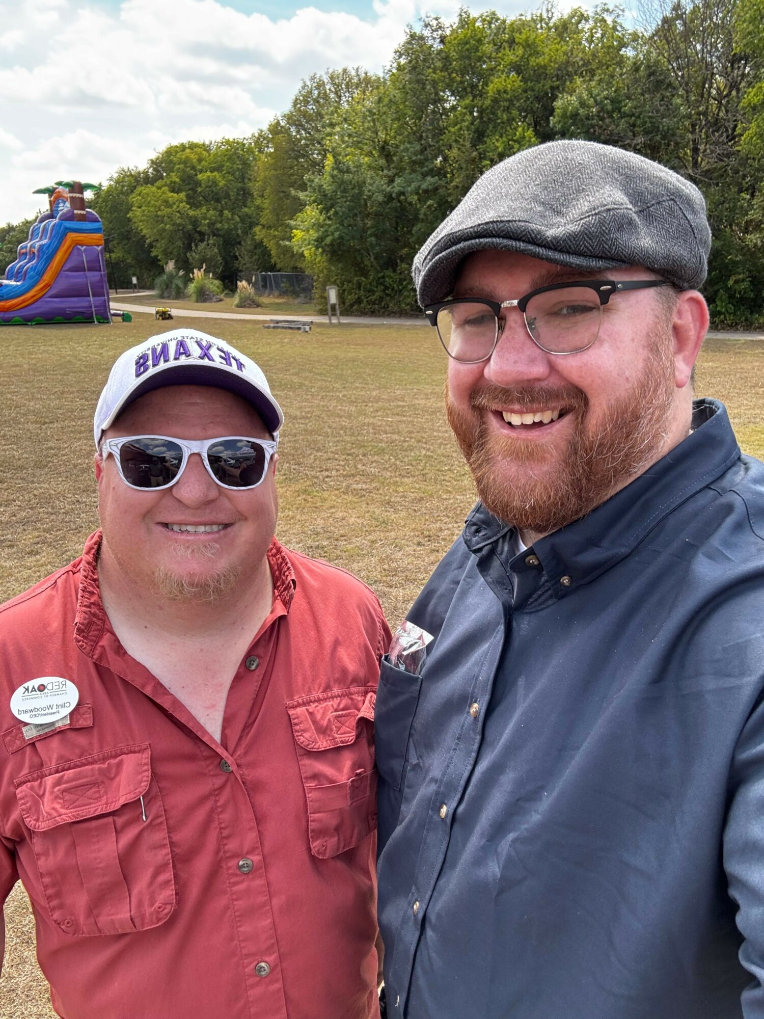 Two men smiling outdoors, one wearing sunglasses and a red shirt, the other wearing a gray cap and glasses, with trees and a bounce house in the background.