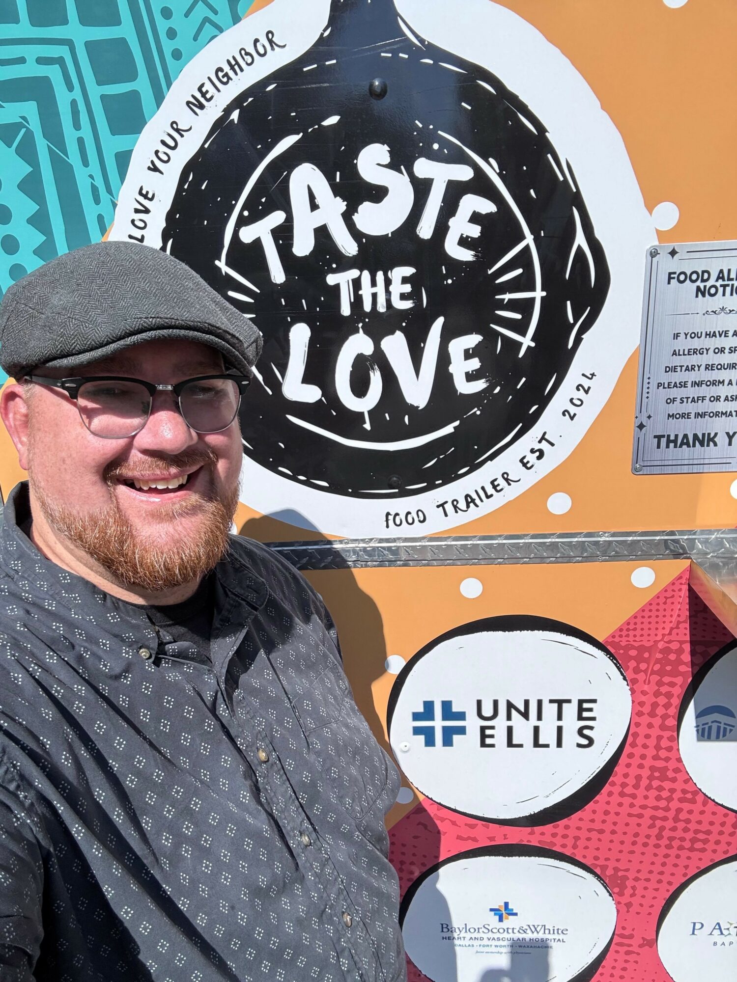 Man with glasses and a gray cap smiling in front of a colorful food trailer sign with the words 