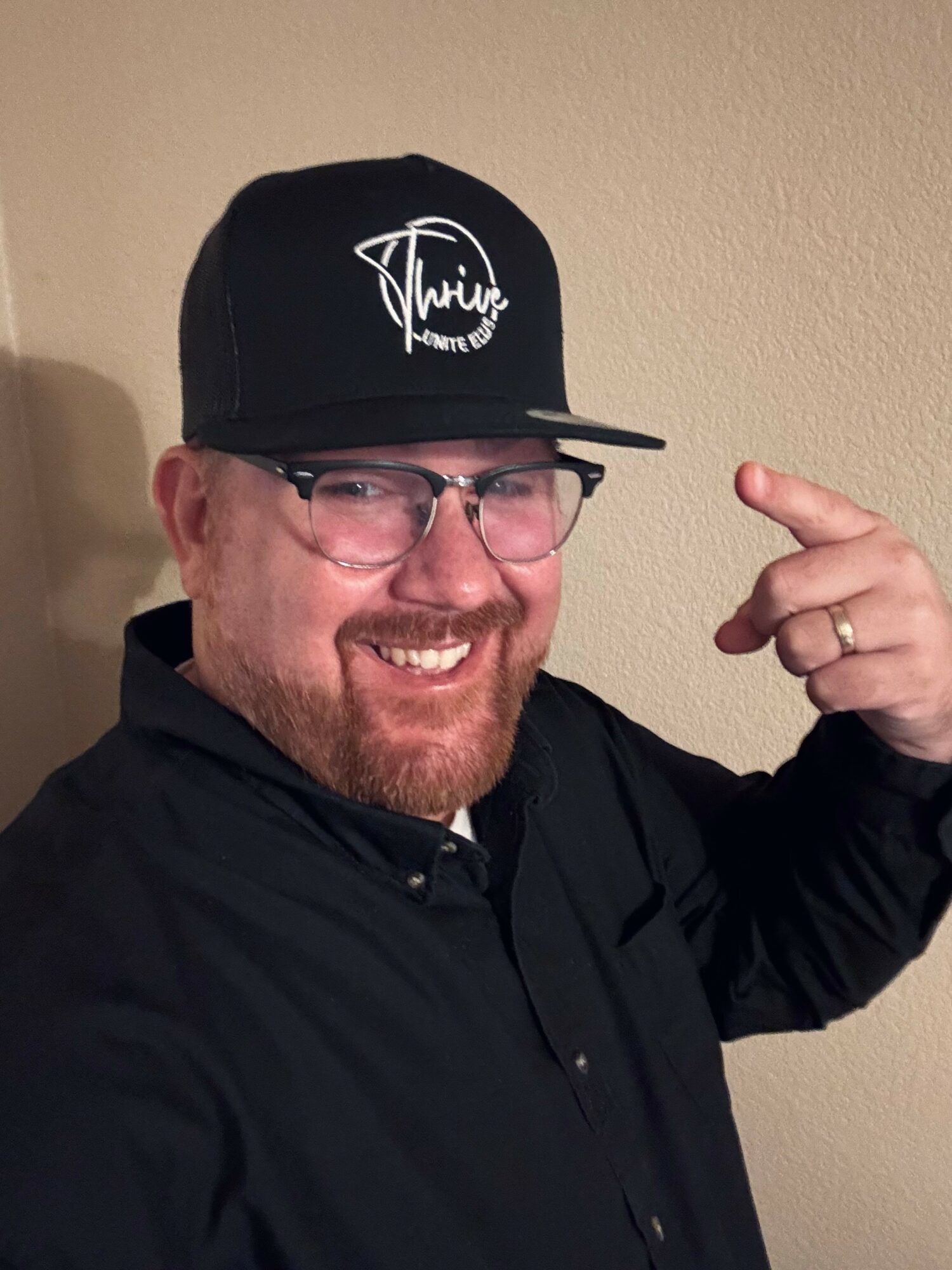 Smiling man wearing glasses and a black cap pointing to his head, against a beige wall.