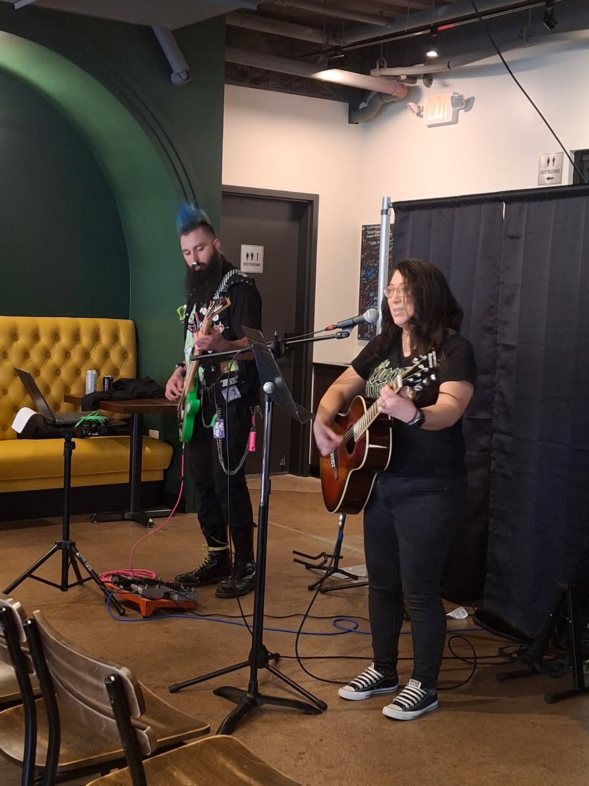 Two women perform with guitars on a stage, one singing into a microphone, in a room with a yellow couch and black curtain.